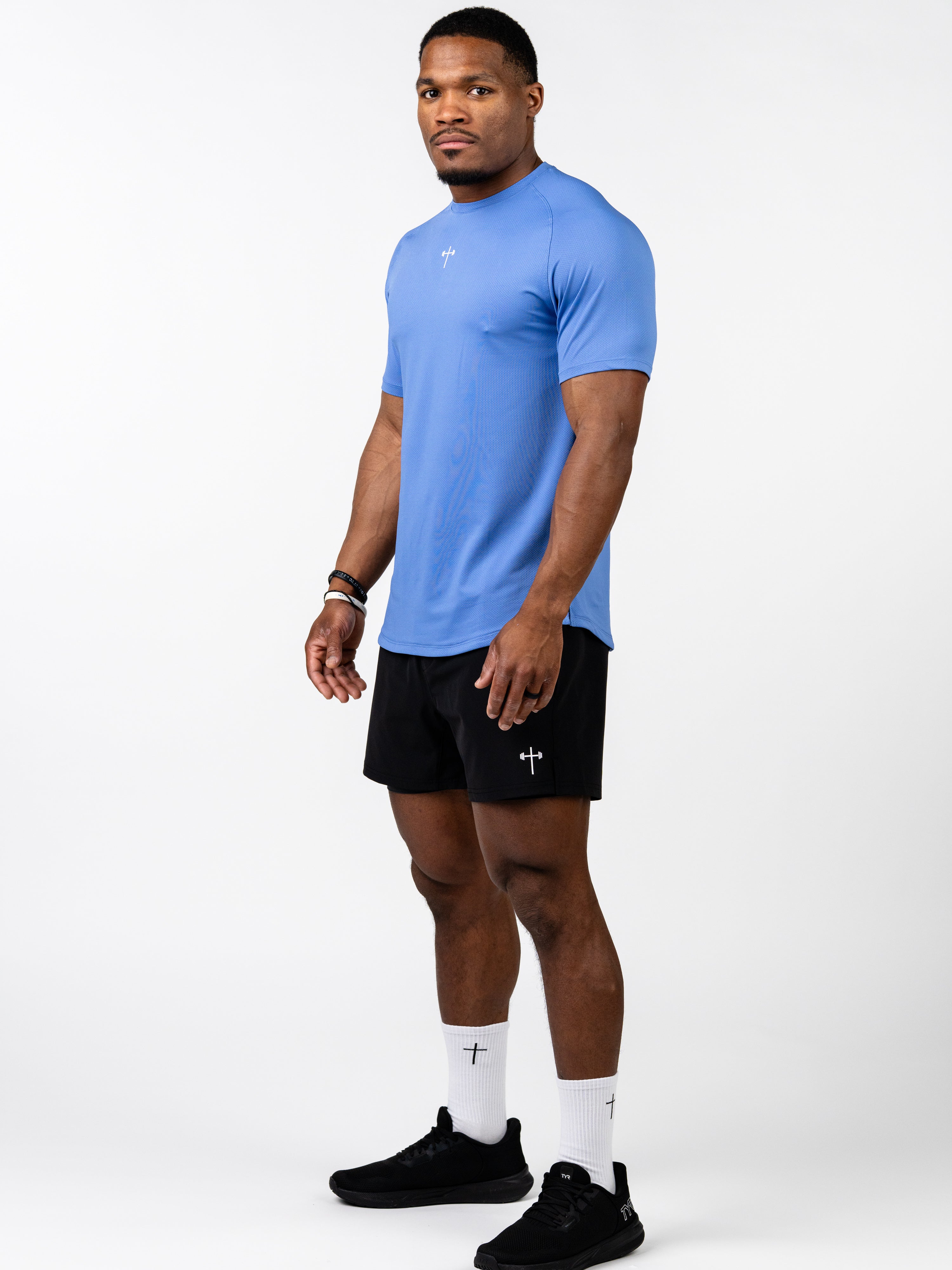Athletic man wearing a blue Signature Tee made of breathable, stretch fabric designed for intense training, paired with black shorts and white socks featuring a Christian cross and barbell symbol.