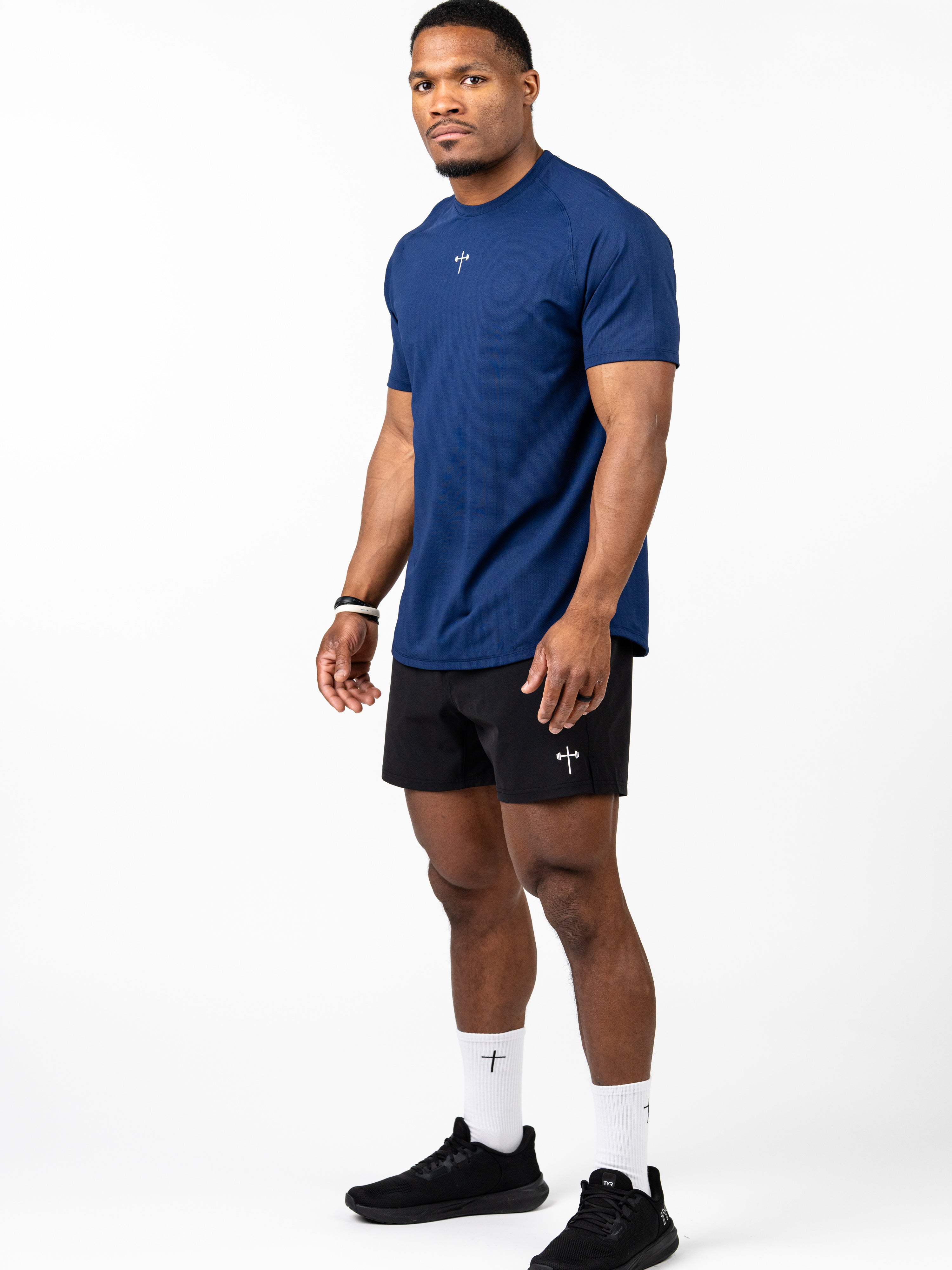Athletic man wearing a navy blue Signature Tee with a small cross and barbell design on the chest, paired with black shorts and white socks featuring a cross, suitable for Christian fitness apparel.