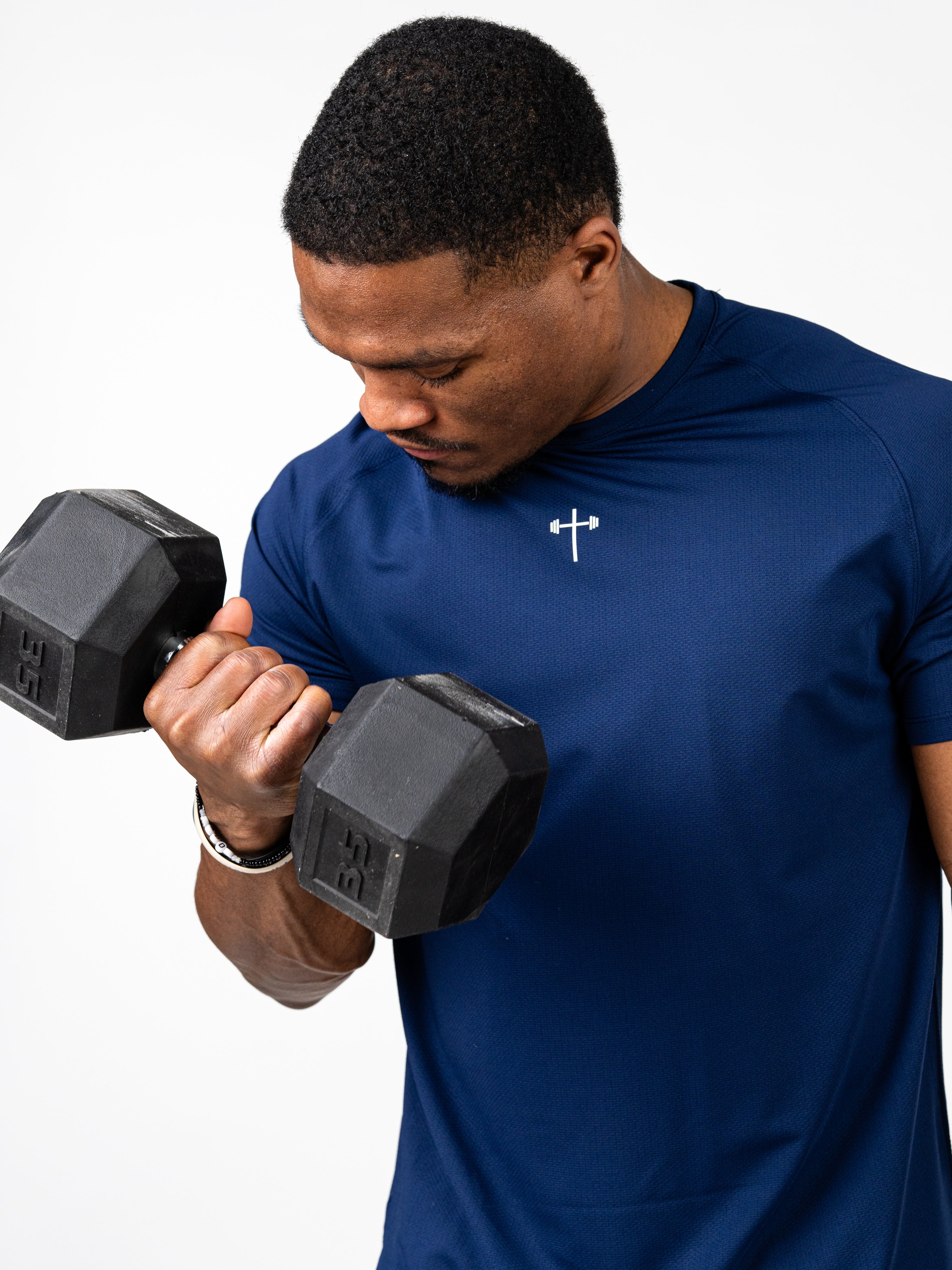 Man wearing navy blue Signature Tee with a small white Christian cross and barbell logo on chest, lifting a black dumbbell during workout.