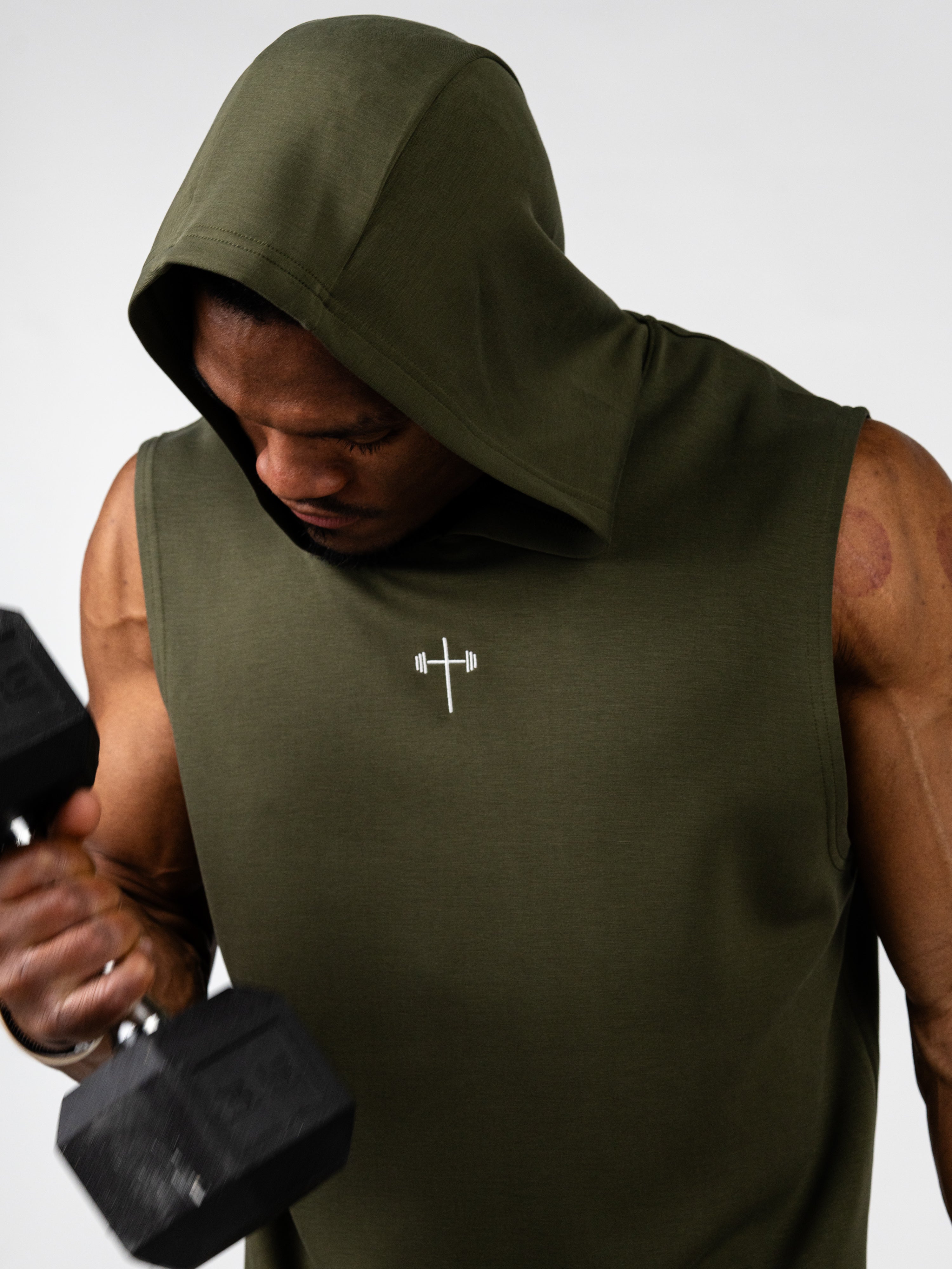 Male athlete wearing an olive green Essential Sleeveless Hoodie with a Christian cross and barbell logo lifting a dumbbell, showcasing faith based fitness apparel.
