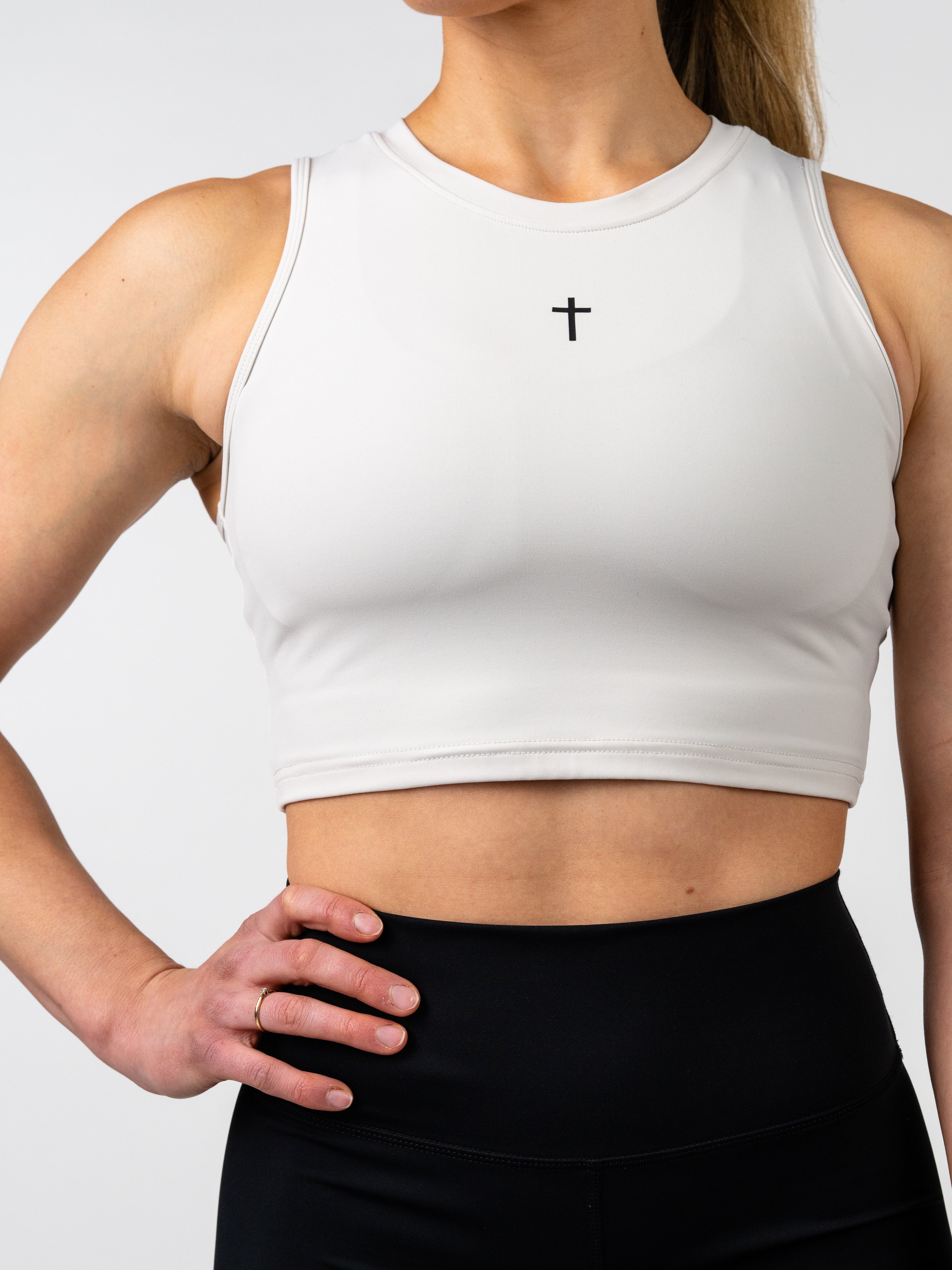Woman wearing a white Vital Cross Combat Top with a small black cross logo centered on the chest, paired with high-waisted black leggings, standing with hand on hip.