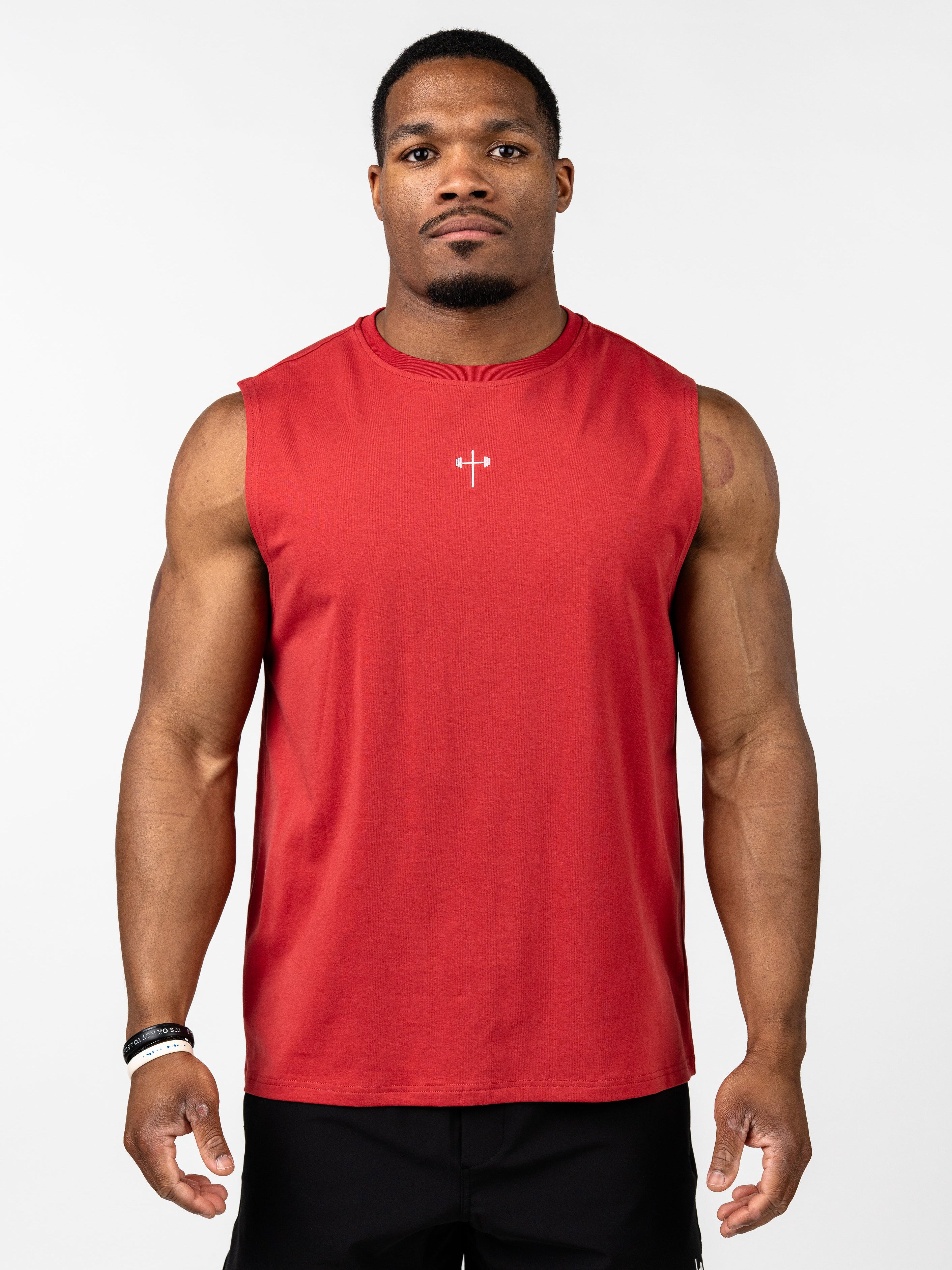 Male model wearing a red Vital Cross Sleeveless Tank designed for high-output training, featuring a small white Christian cross with a barbell emblem on the chest, paired with black shorts.