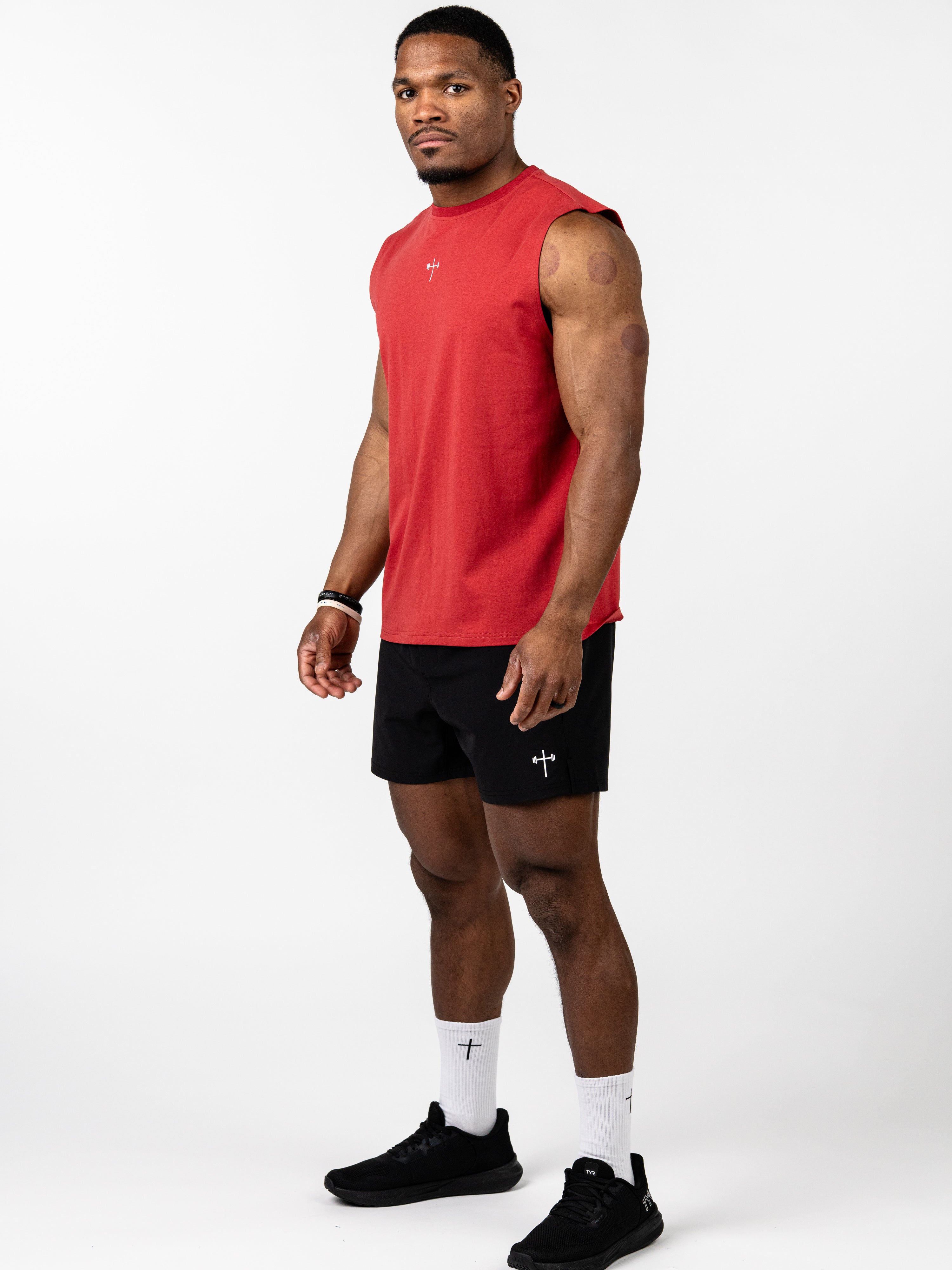 Athletic male model wearing a red Vital Cross Sleeveless Tank with a small cross emblem, paired with black shorts and white socks featuring Christian gym clothing symbols.