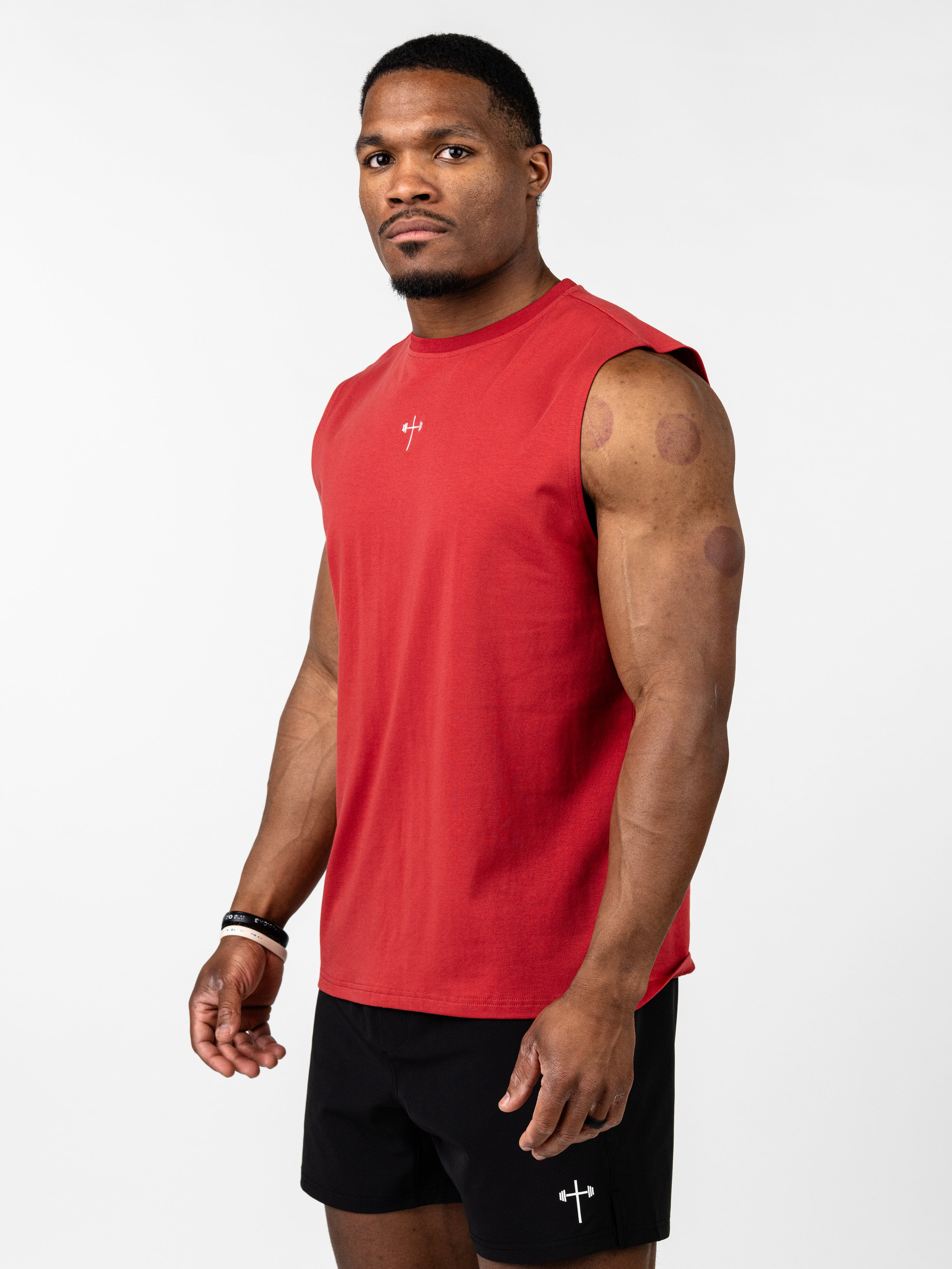 Man wearing a red Vital Cross Sleeveless Tank with a small Christian cross and barbell design on the chest, paired with black shorts featuring a matching logo, standing against a white background.