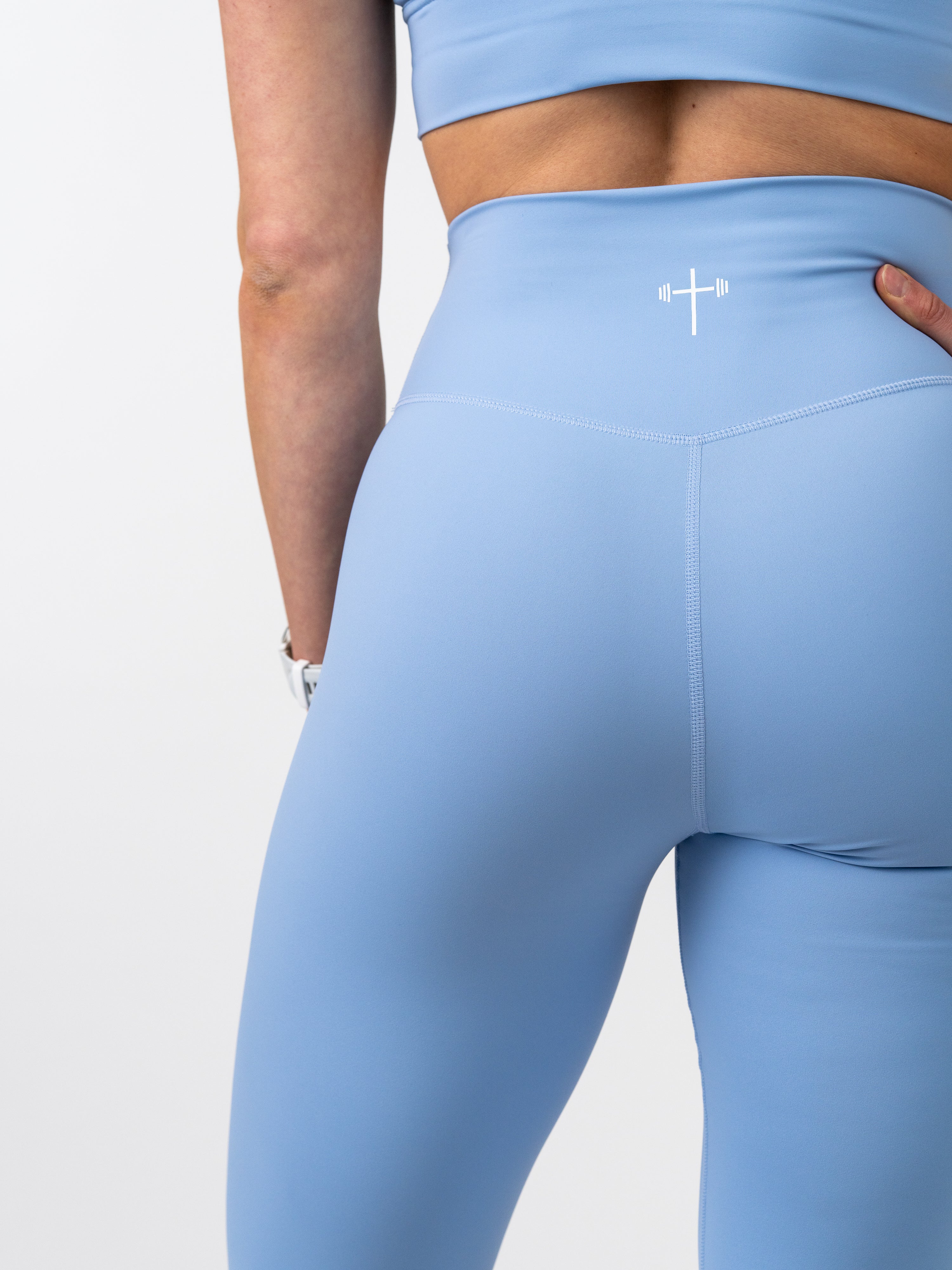 Close-up of a person wearing light blue Essential Leggings featuring a high waistband and a small white Christian cross with a barbell logo on the back, designed for faith-based fitness.