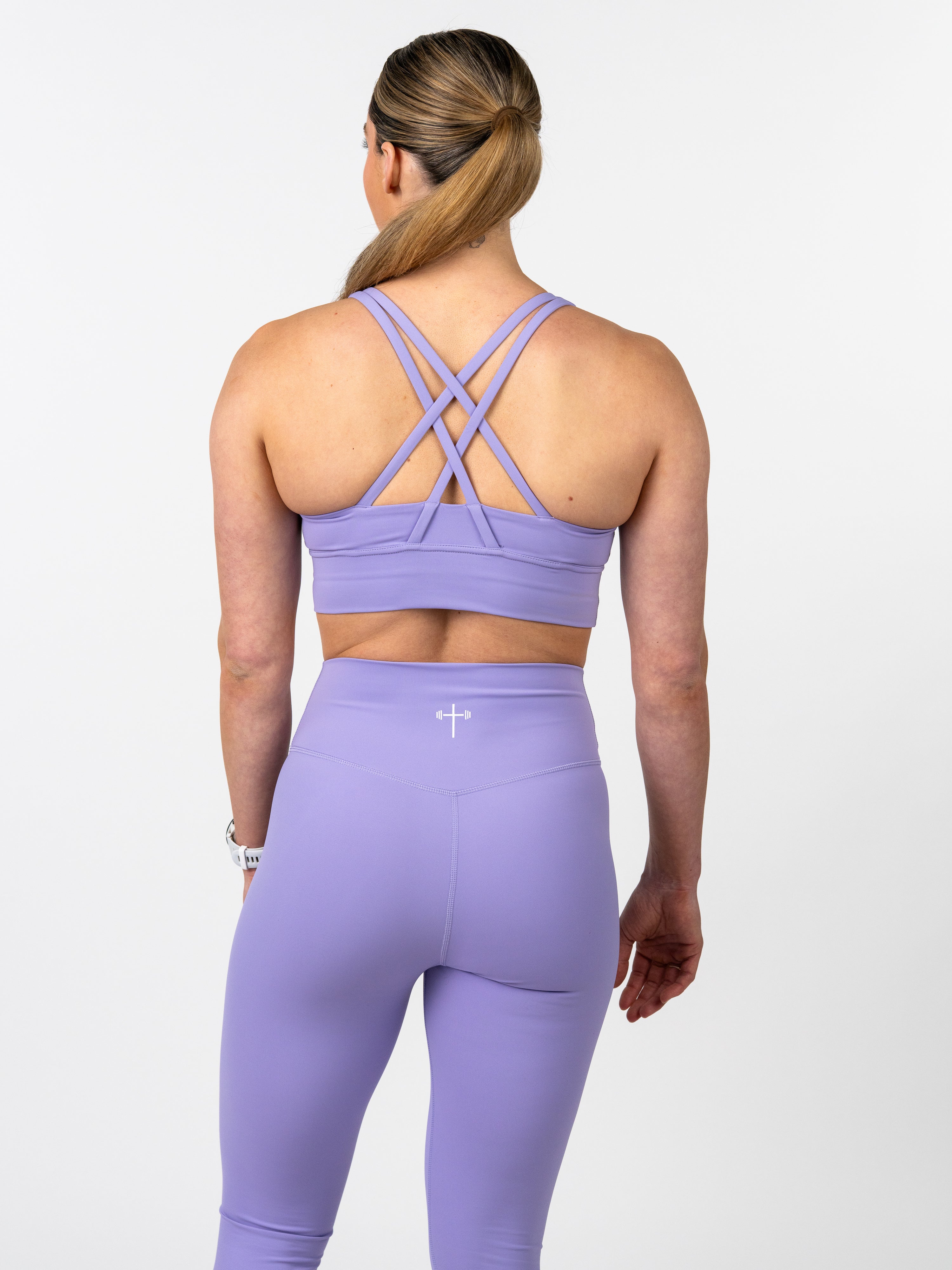 Woman wearing a lavender Essential Sports Bra with a unique cross back strap design, paired with matching high-waisted leggings featuring a subtle cross and barbell logo.