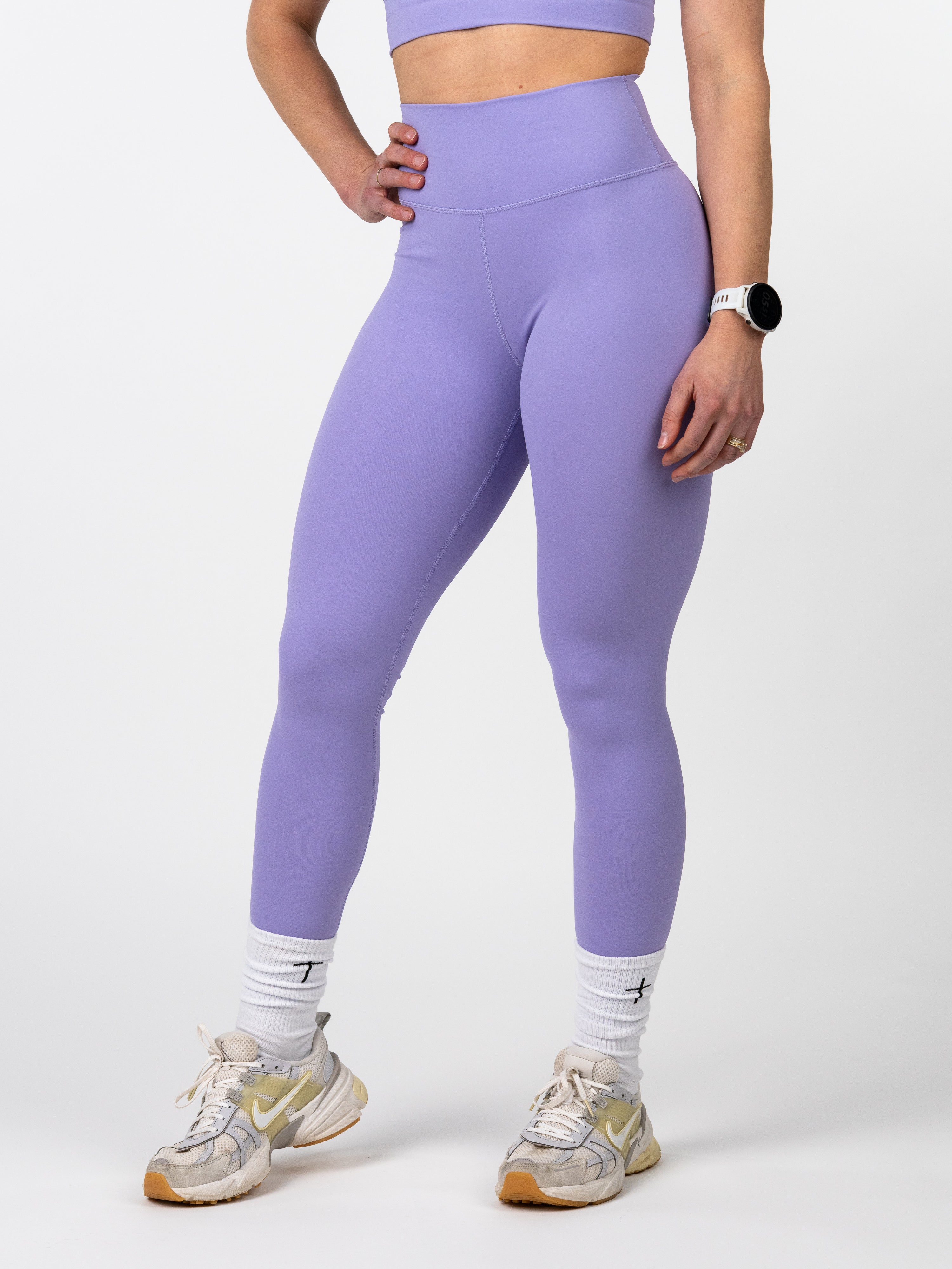 Woman wearing purple Essential Leggings designed for endurance and flexibility, paired with white athletic socks and running shoes, standing against a white background.