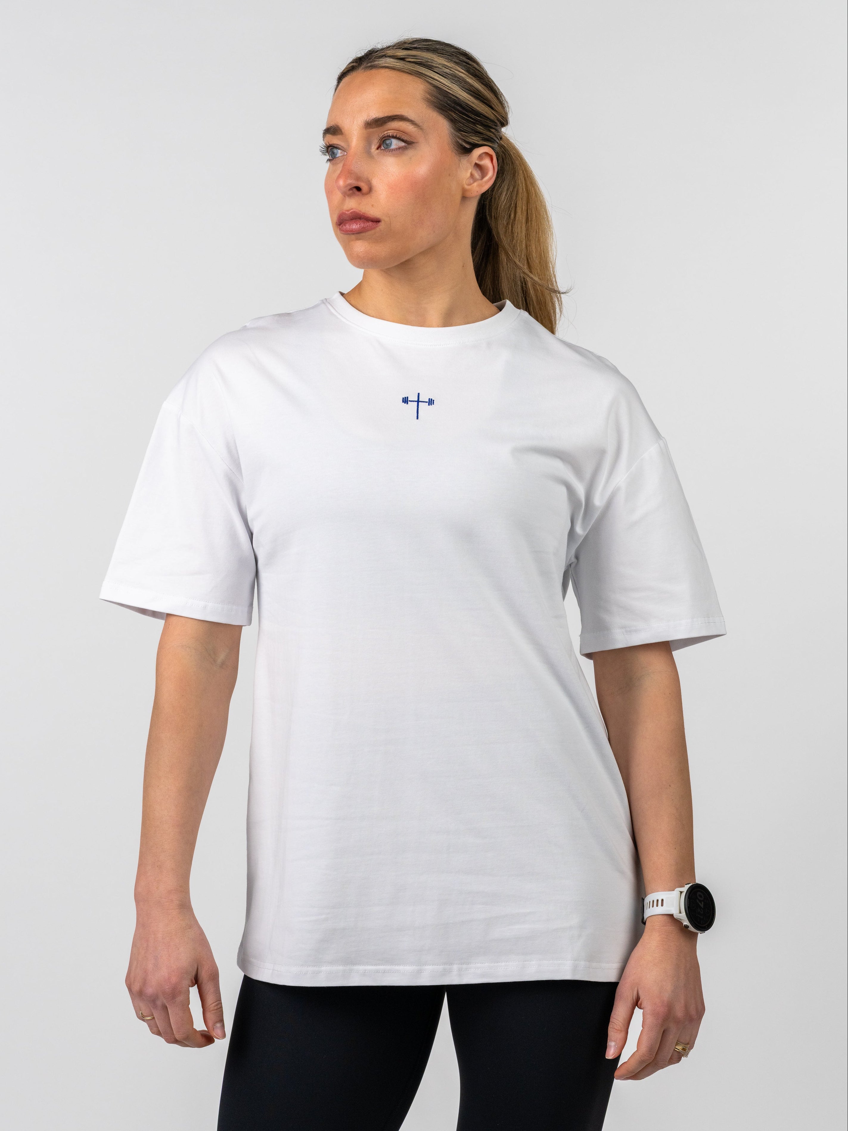 Woman wearing a white Women's Lifting Club Oversized Tee featuring a small blue cross and barbell graphic on the chest, styled with black leggings against a plain background.