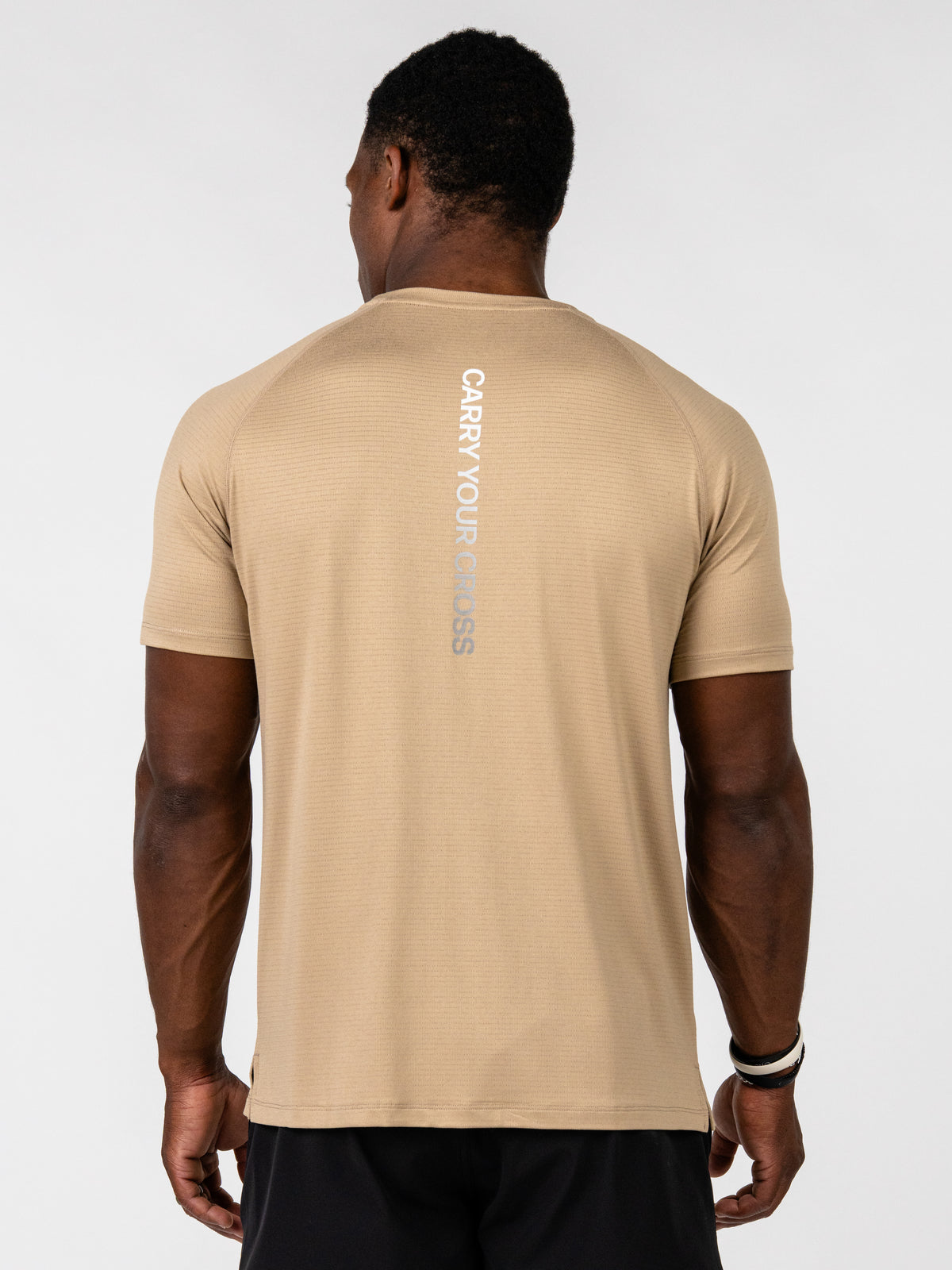 A man viewed from the back wearing a beige athletic t-shirt with 'CARRY YOUR CROSS' printed vertically in gray on the back, paired with black shorts against a light gray studio background.