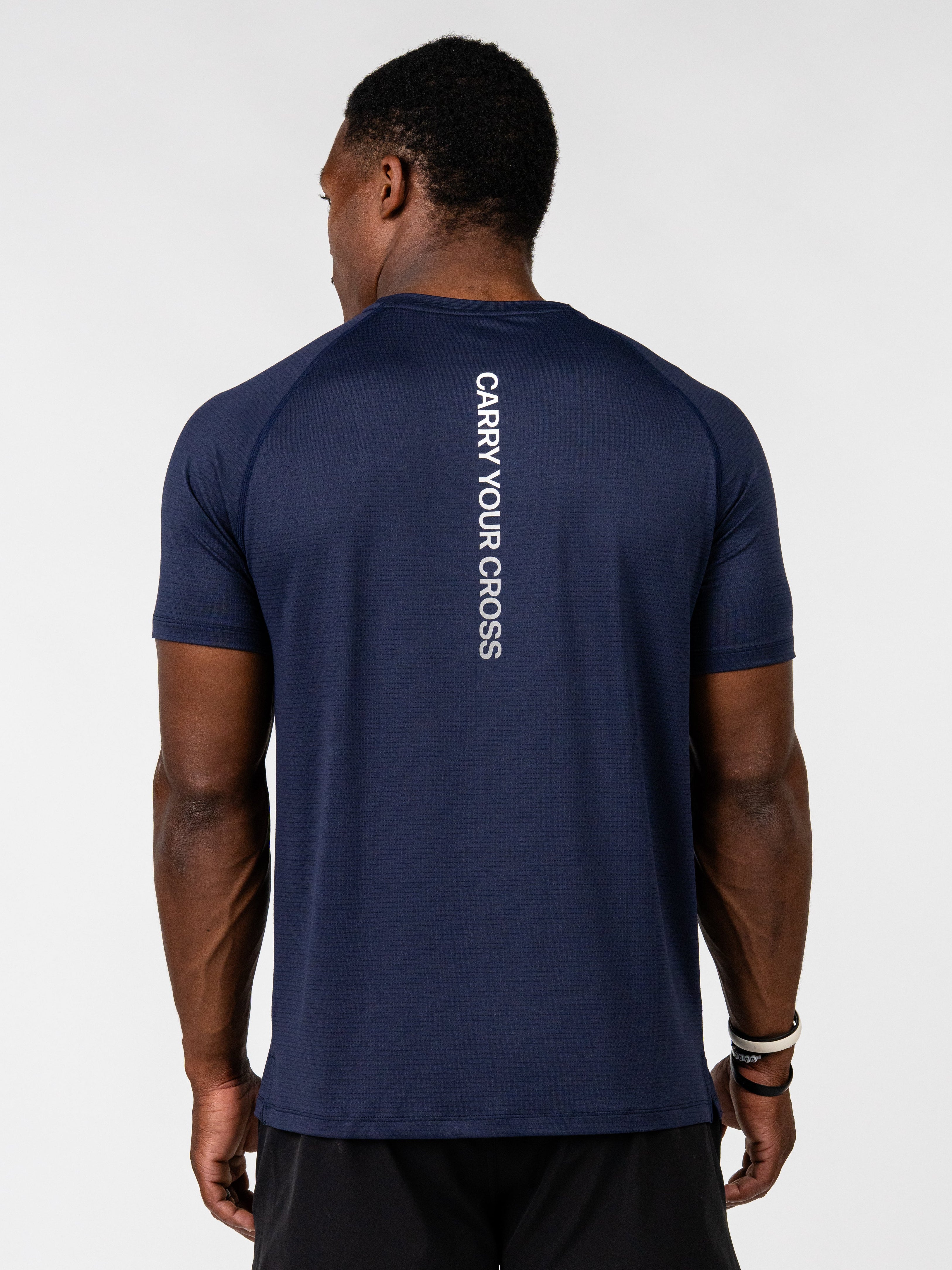 A man viewed from behind wearing a navy blue athletic t-shirt with 'CARRY YOUR CROSS' printed vertically in white on the back, paired with black athletic shorts, against a light gray studio background.