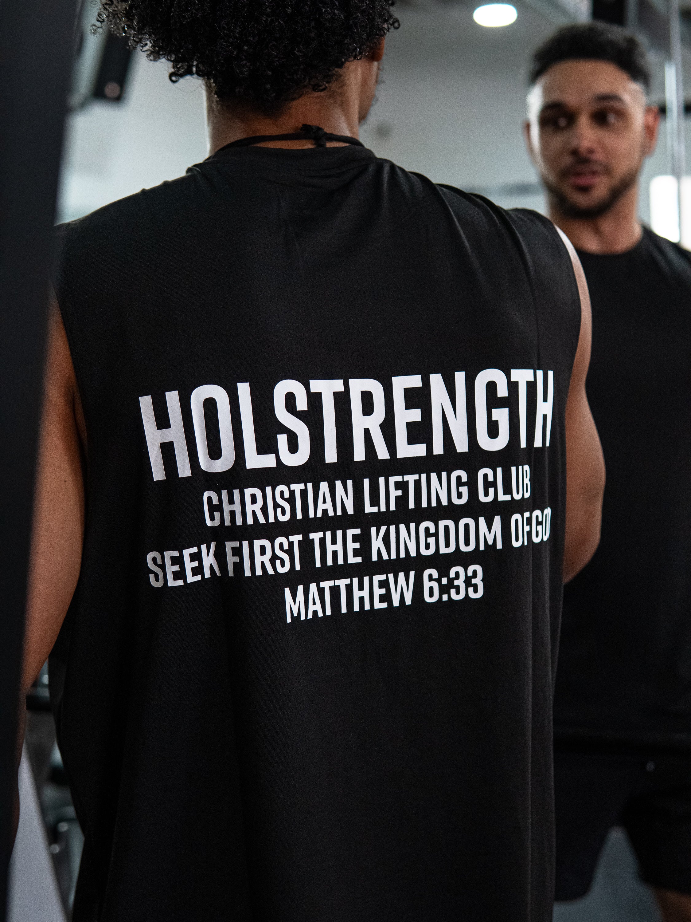 Rear view of a person wearing the Christian Lifting Club Premium Oversized Tank in black with white text that reads "HOLSTRENGTH Christian Lifting Club Seek First the Kingdom of God Matthew 6:33" during a gym workout session.
