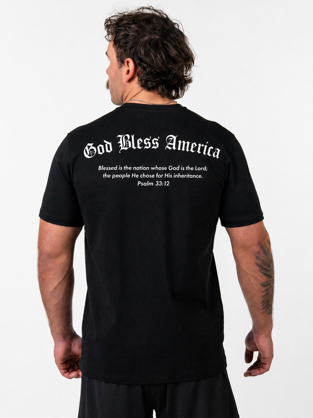 A man viewed from the back wearing a black t-shirt with white gothic text that reads 'God Bless America' and a Bible verse printed on the back.