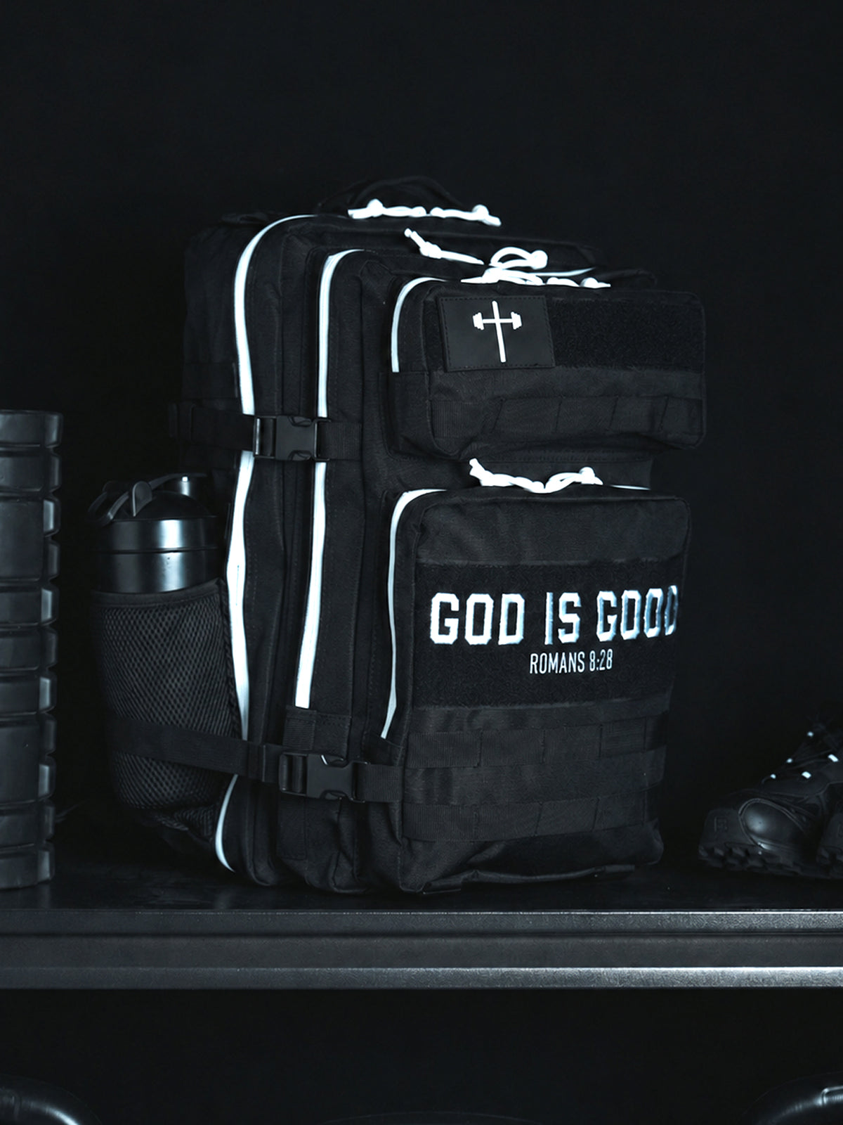 HolStrength 45L Tactical Backpack in black with multiple compartments white trim and God Is Good Romans 1:23 lettering displayed in a gym setting