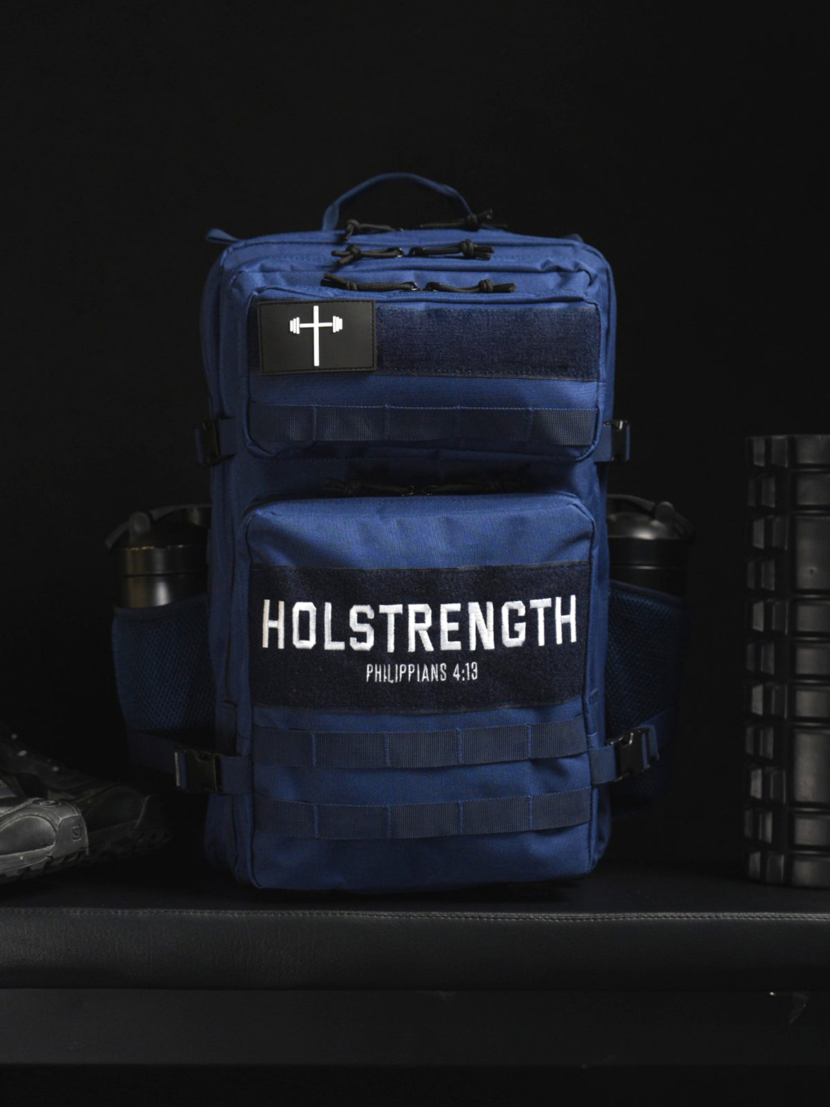 HolStrength 45L Tactical Backpack in navy blue with multiple zippered compartments MOLLE webbing and front cross patch displayed in a gym setting