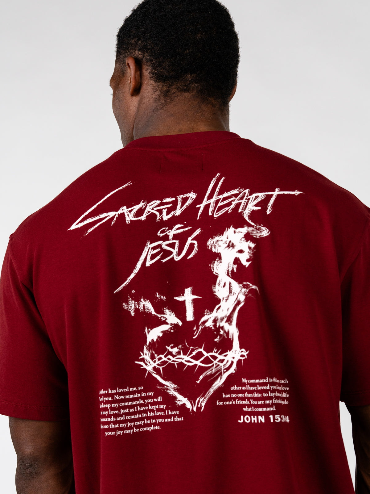 Sacred Heart Premium Oversized Tee shown from the back in red with a white Sacred Heart of Jesus graphic, cross, scripture text, and John 15 34 reference