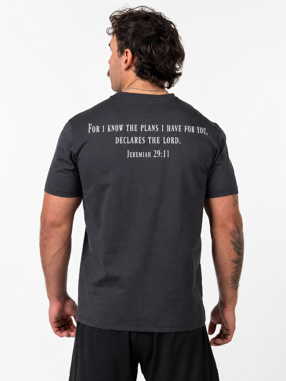 A man viewed from the back wearing a dark charcoal gray t-shirt with a white printed Bible verse from Jeremiah 29:11 on the upper back, paired with black athletic shorts, against a plain white background.