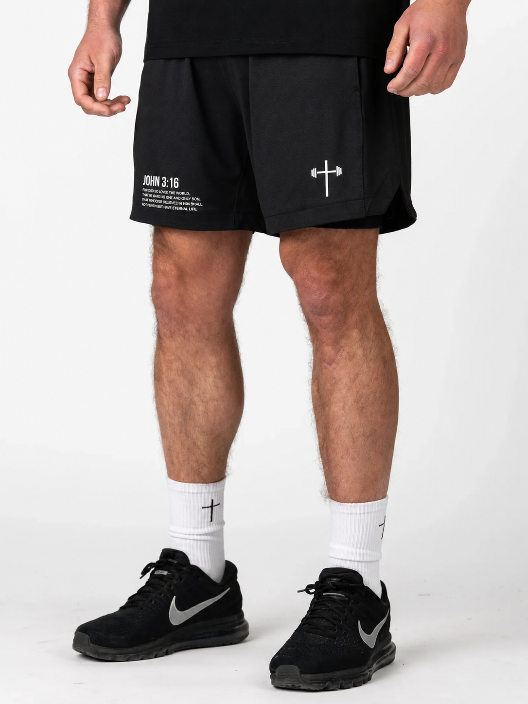 Person wearing black shorts with a white logo, white socks, and black sneakers on a white background. Shorts say John 3:16