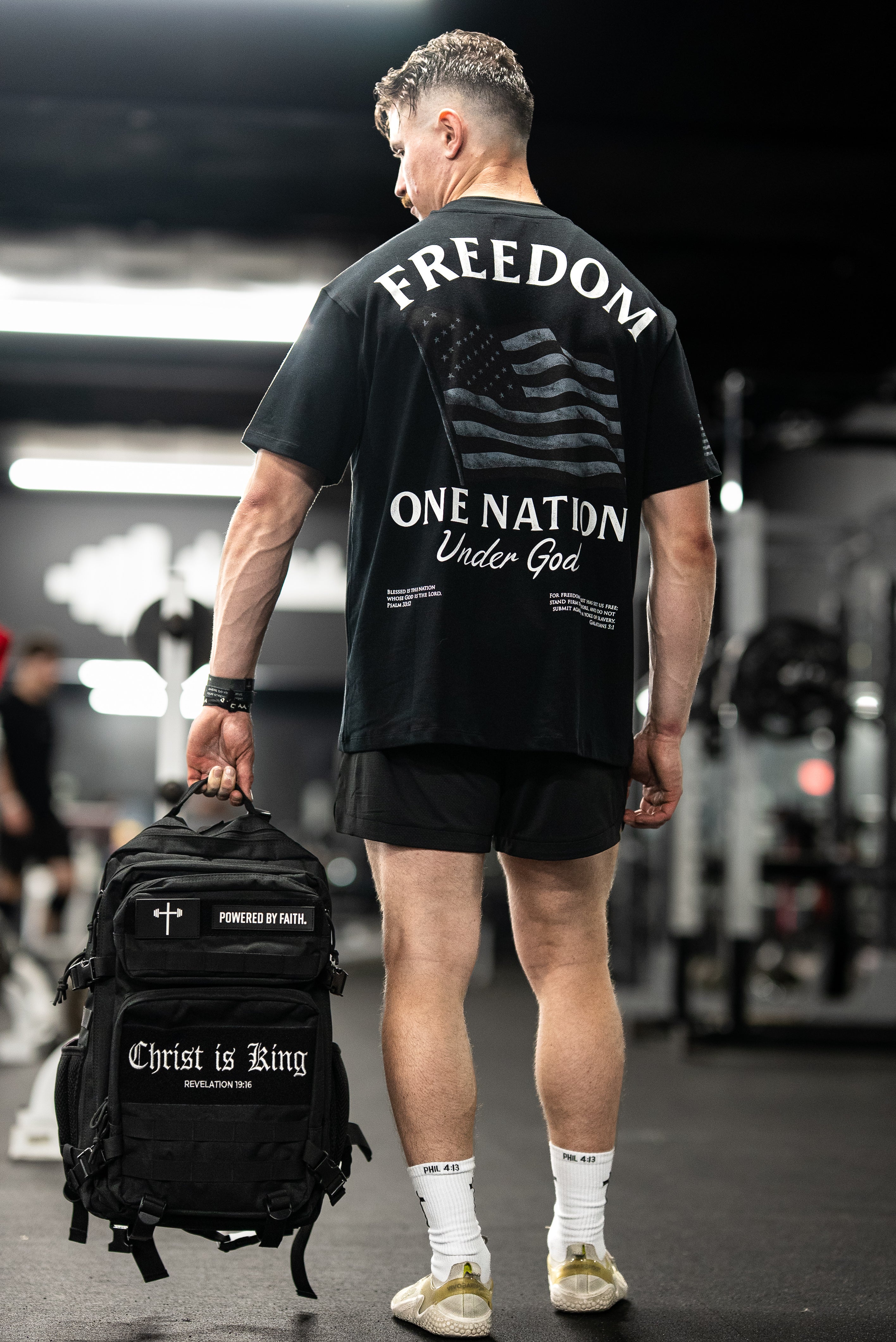 Man wearing a black One Nation Under God Oversized Tee with a faded American flag and bold "FREEDOM" text, holding a black backpack with faith-based messages in a gym setting.
