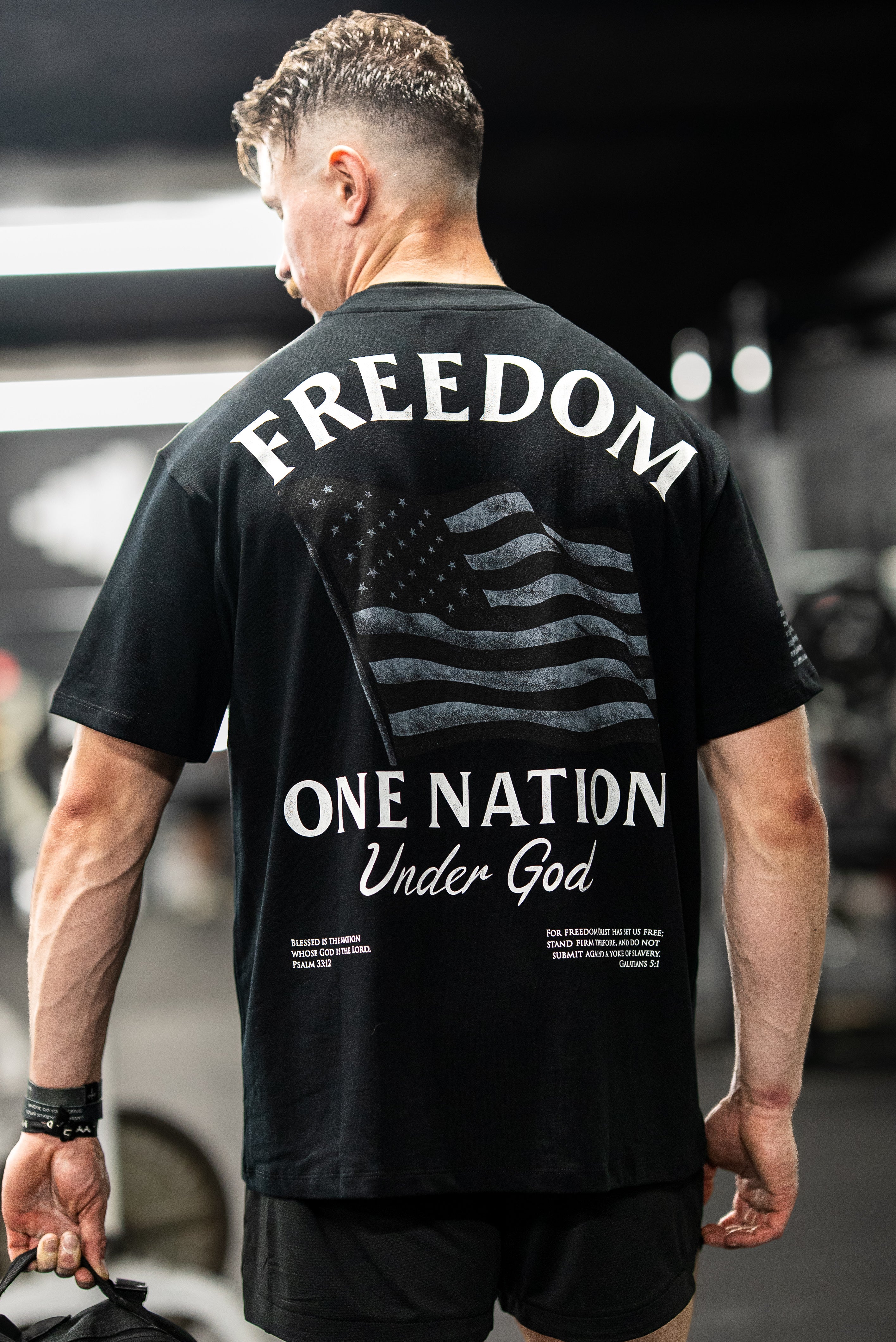 Man wearing a black One Nation Under God Oversized Tee with a large faded American flag and faith-based text print on the back, shown in a gym setting.