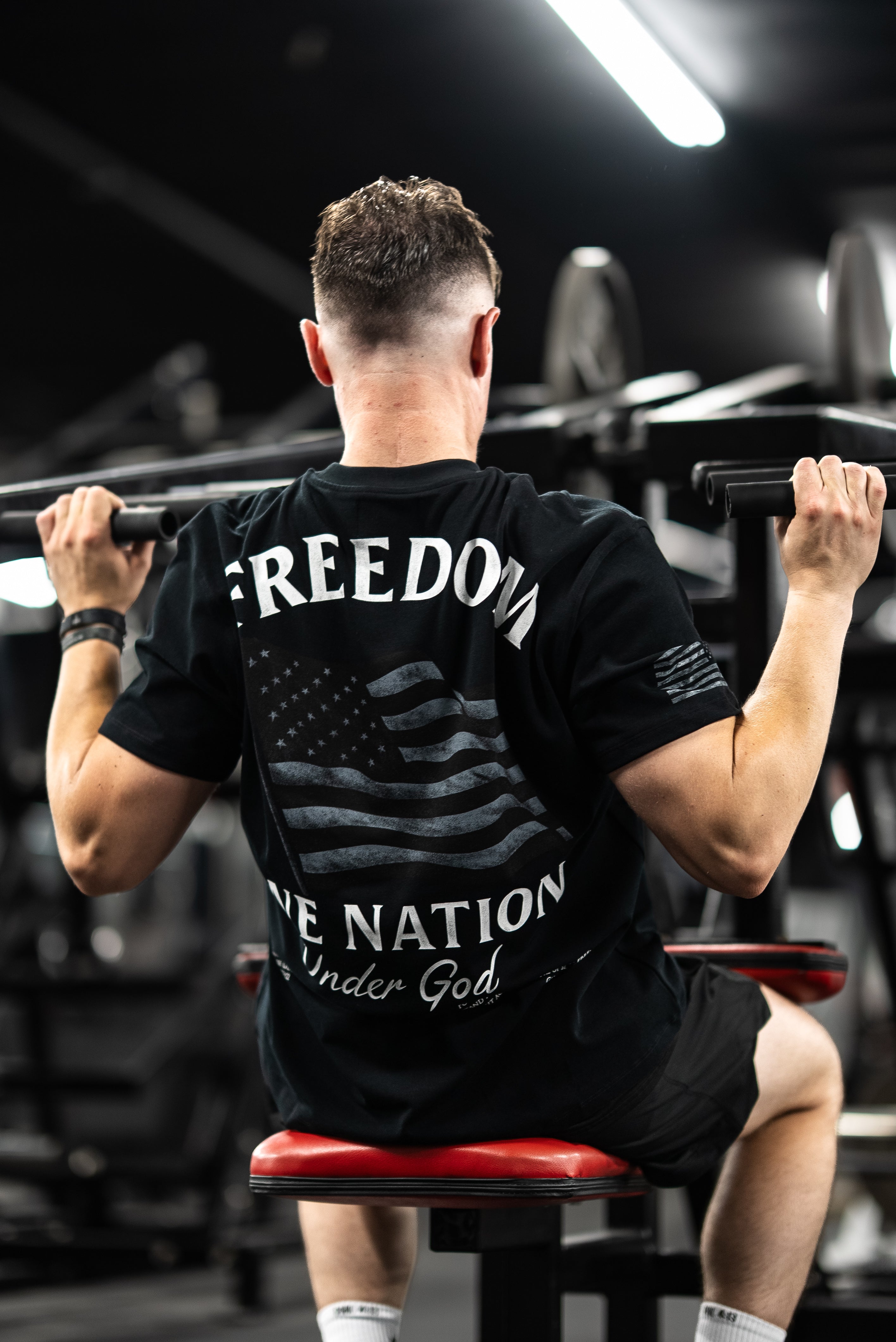 Man lifting weights at gym wearing a One Nation Under God Oversized Tee with a black and white American flag design promoting faith and discipline in Christian fitness apparel