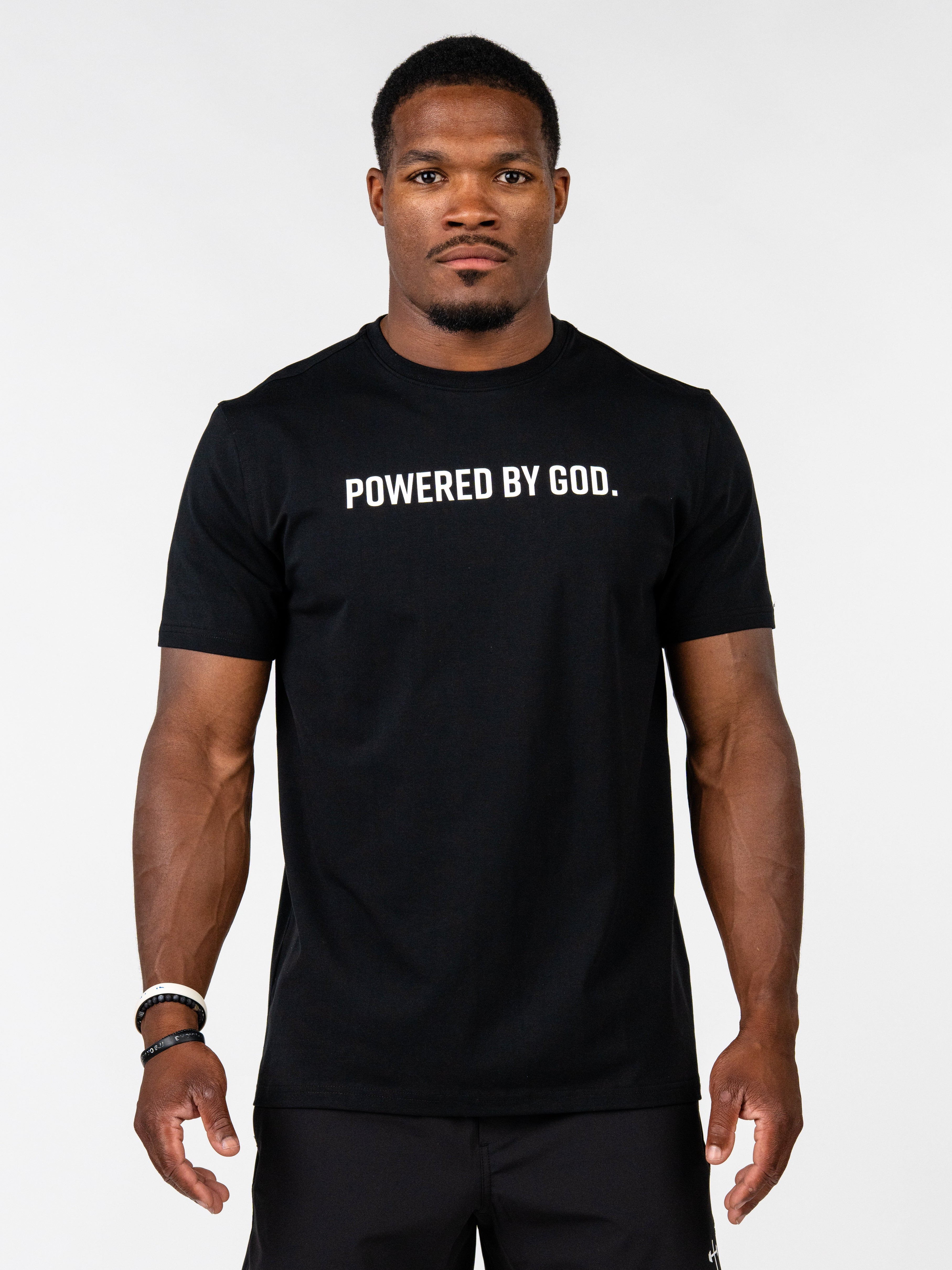 A man wearing a black short-sleeve t-shirt with the white printed text 'POWERED BY GOD.' across the chest, standing against a light gray studio background.