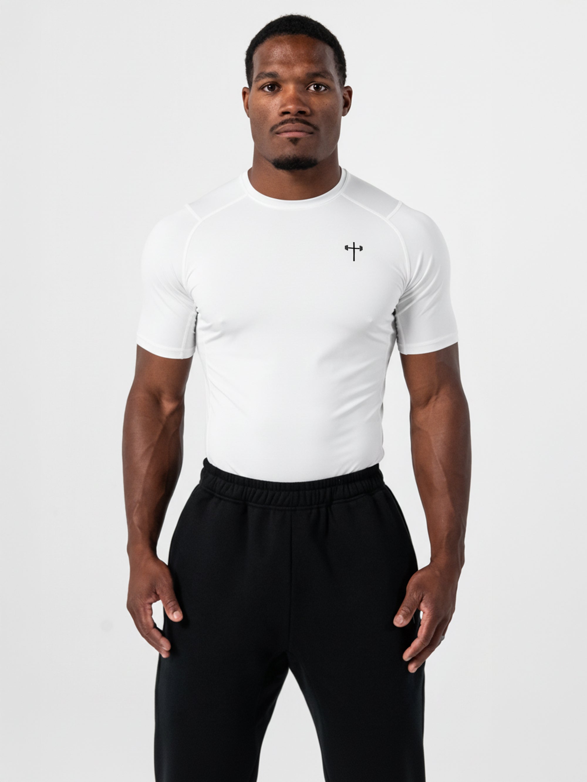 Athletic man wearing a white Core Base Layer Tee with a lightweight compression fit and a small cross logo on the chest, paired with black workout pants.