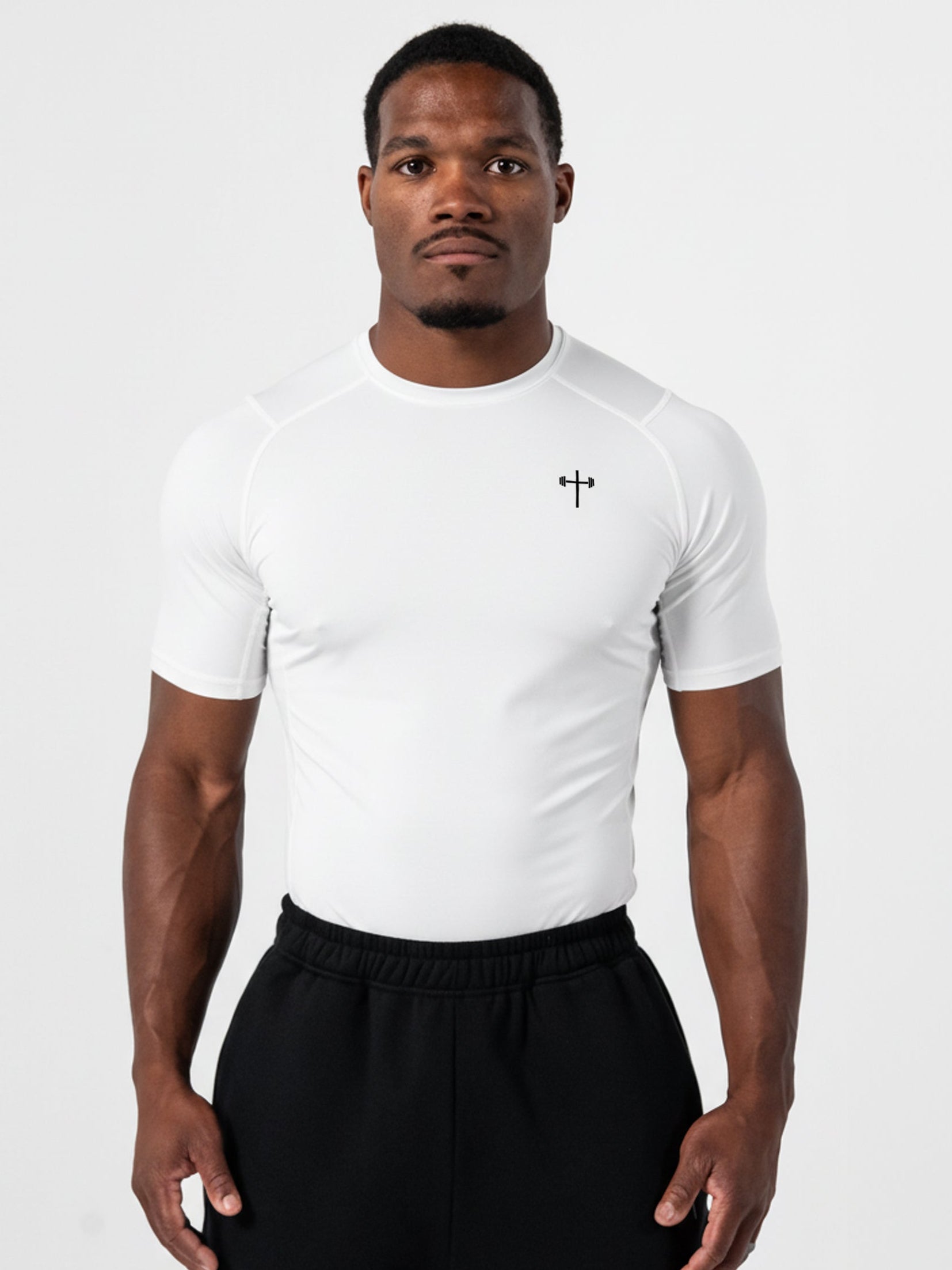 Athletic man wearing a white Core Base Layer Tee with a lightweight compression fit and a small cross logo on the chest, paired with black workout pants.