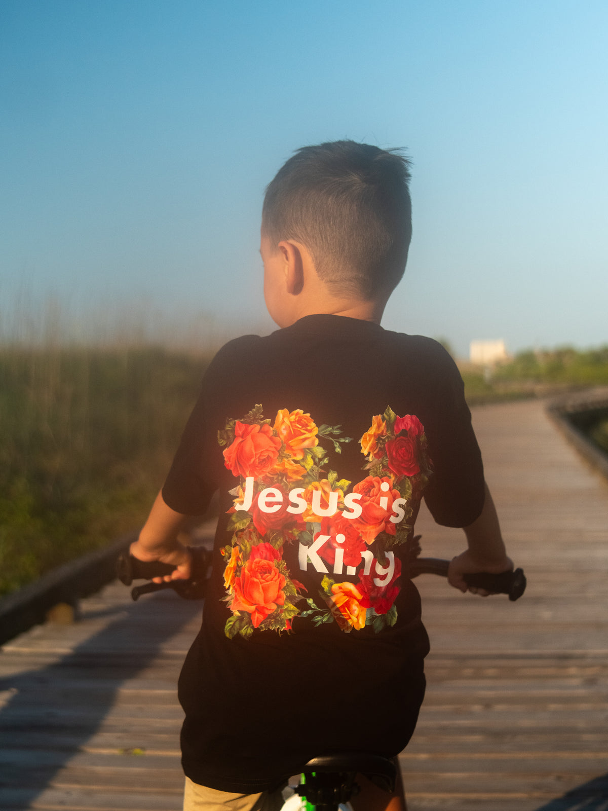 Child riding a bicycle outdoors wearing a black Jesus is King Youth Tee featuring colorful floral graphics and bold text on the back, under clear blue sky.