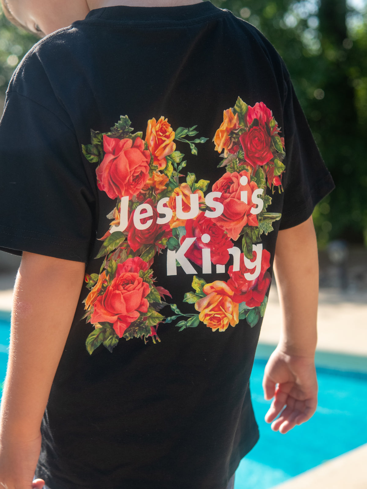 Back view of a youth wearing a black Jesus is King Youth Tee featuring a colorful floral graphic with red and orange roses around the text near a bright outdoor pool setting.