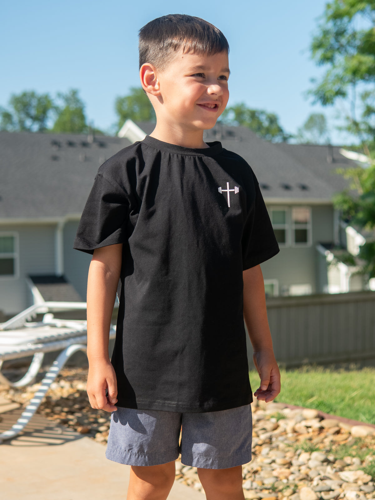 Young boy standing outdoors wearing a black Jesus is King Youth Tee featuring a small white cross logo on the front, paired with gray shorts on a sunny day.