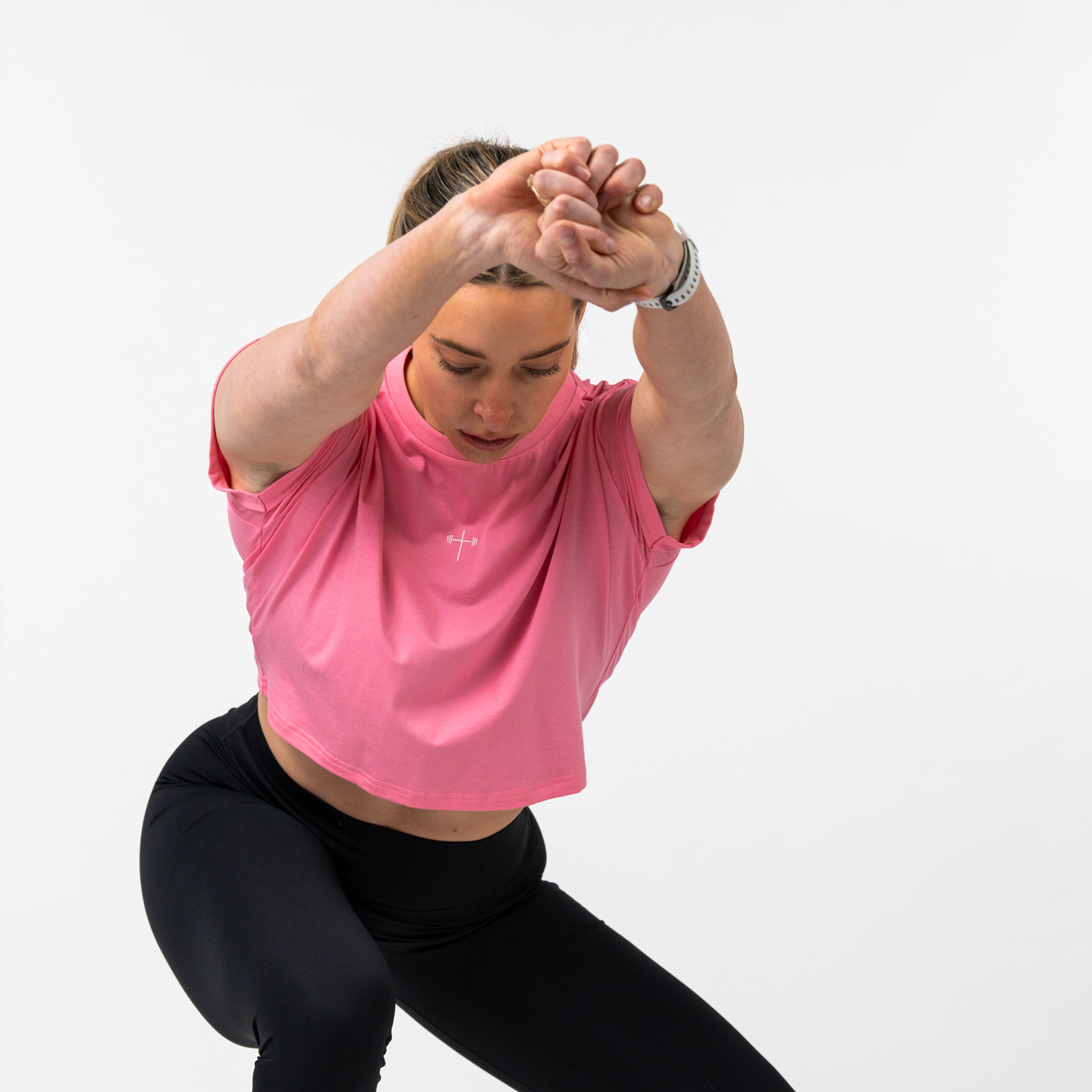 Woman wearing a pink cropped t-shirt with a small cross and barbell graphic on the chest, stretching her arms forward while wearing black leggings against a plain white background.