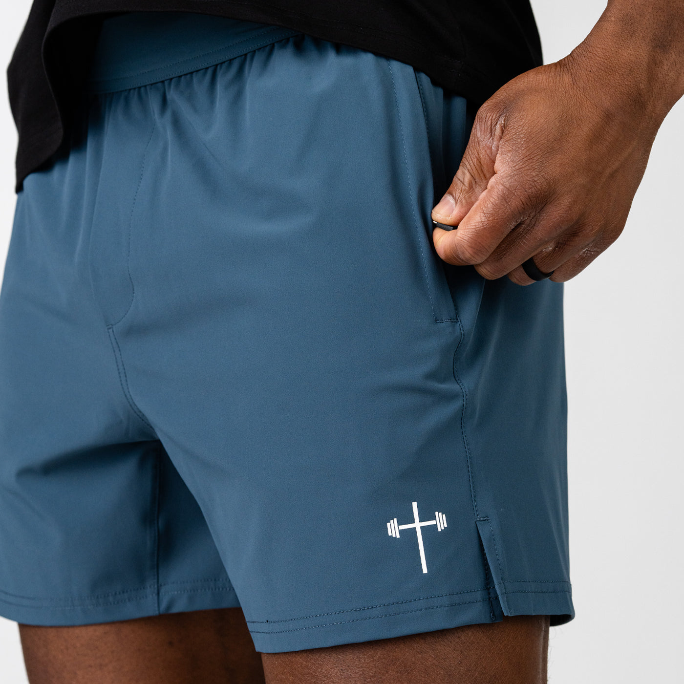 Close-up of a person wearing dark blue athletic shorts featuring a small white barbell and cross logo near the hem, with a hand resting near the side pocket.