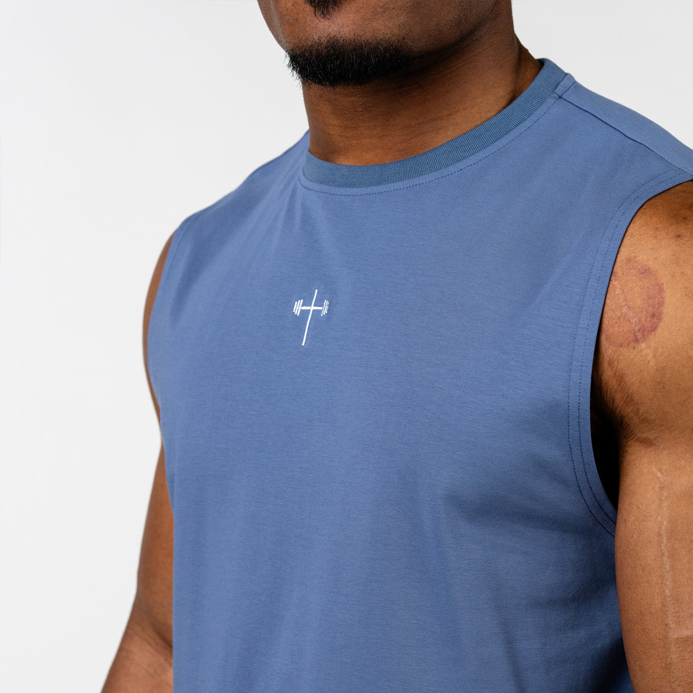 Close-up of a man wearing a blue sleeveless workout shirt featuring a small white barbell cross emblem centered on the chest, highlighting Christian fitness apparel.