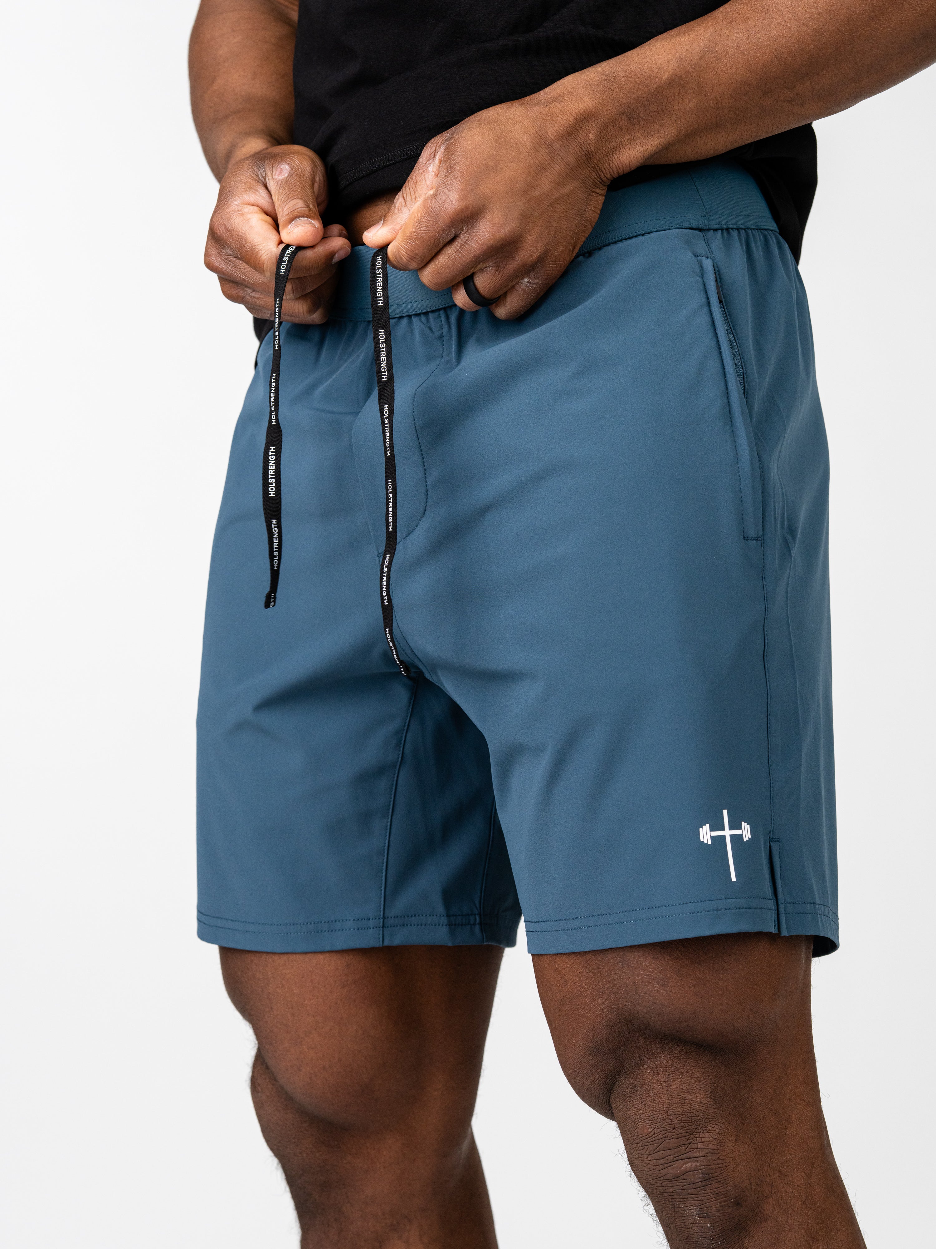 Man adjusting the drawstring on blue Essential Training Shorts with a small white cross and barbell logo on the left leg, made for lightweight, breathable comfort during workouts.