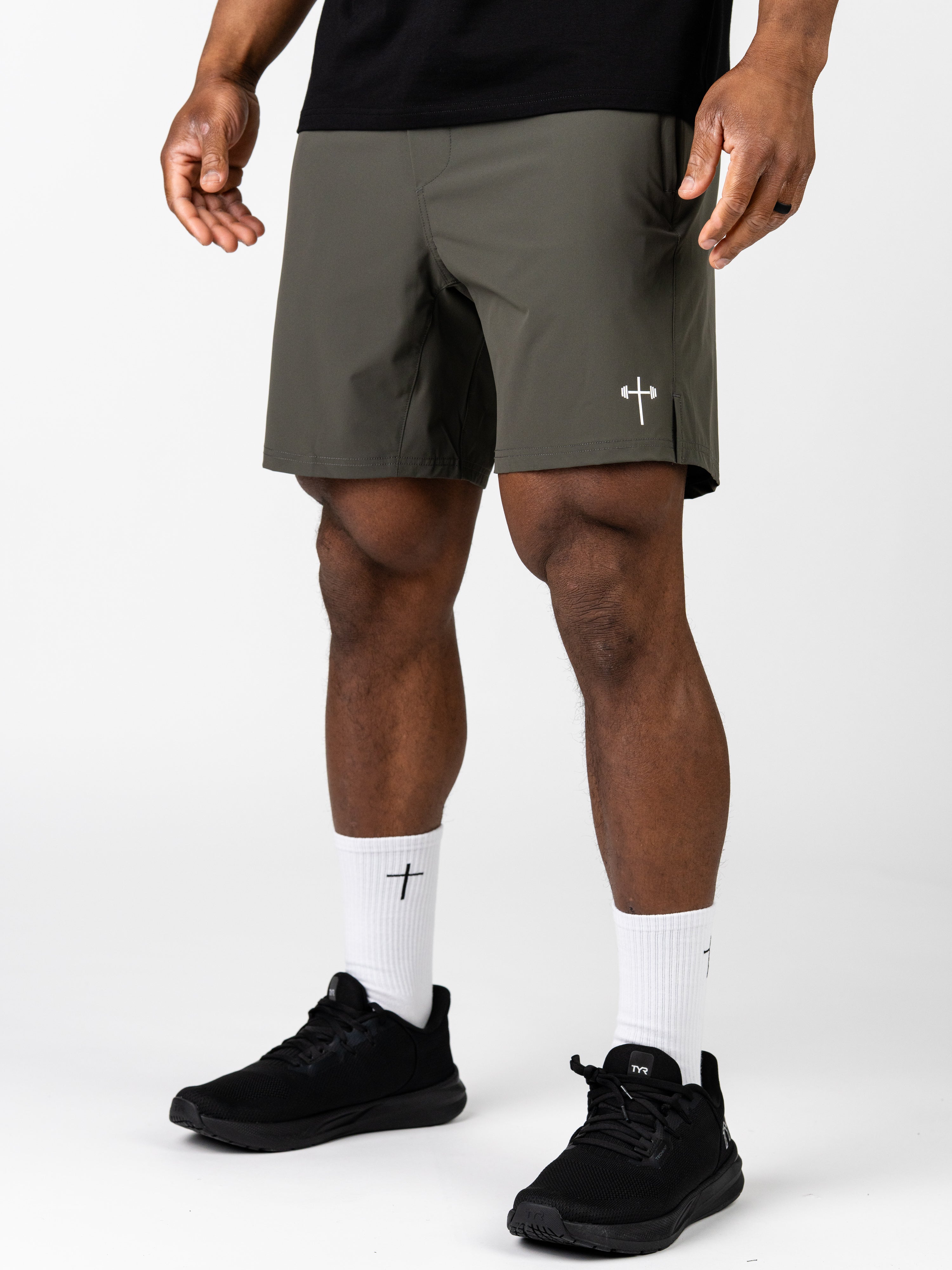 Man wearing olive green Essential Training Shorts with a small white cross and barbell logo on the leg, paired with white socks featuring a black cross and black athletic shoes.