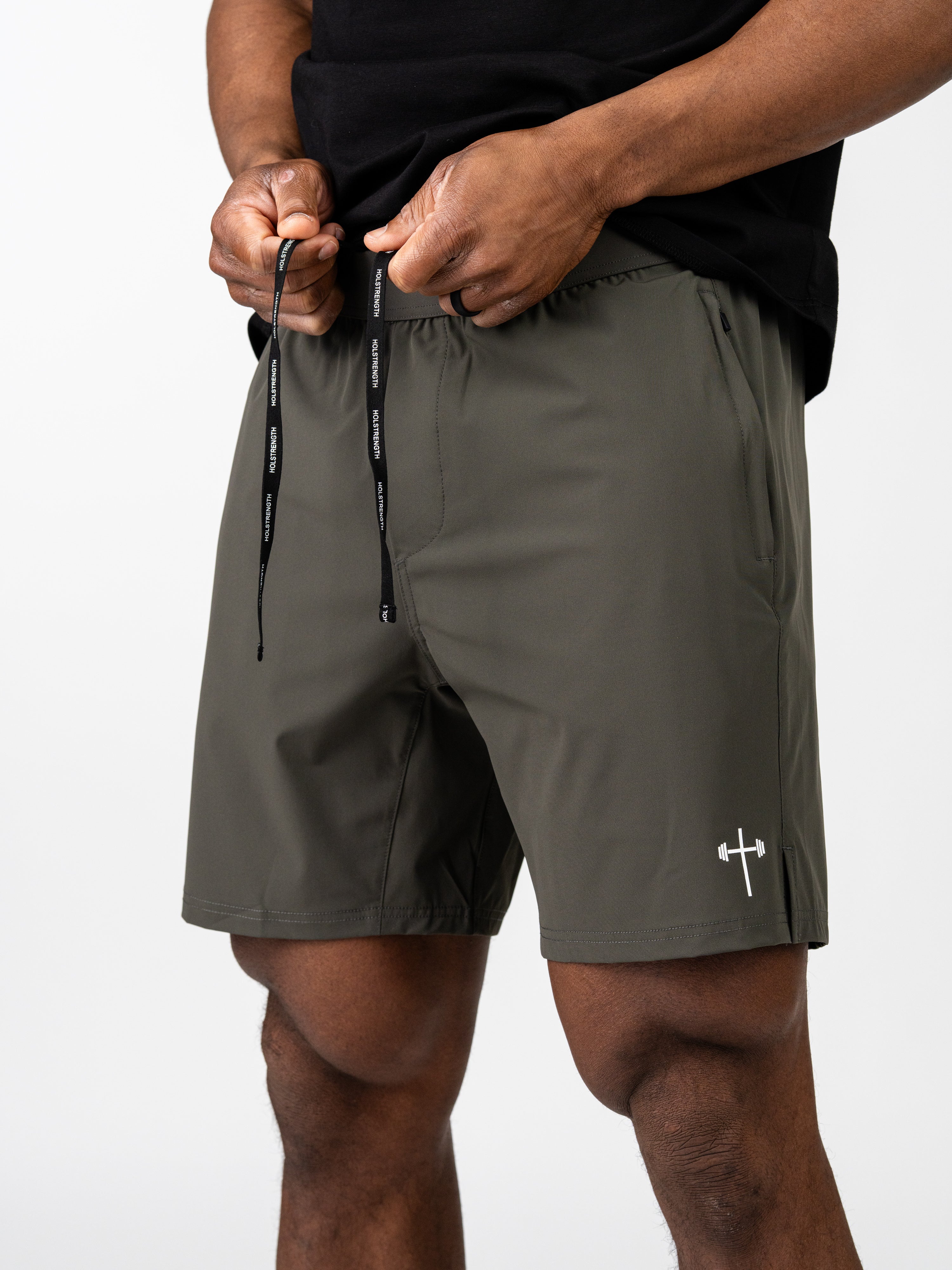 Close-up of a person adjusting the drawstring on olive green Essential Training Shorts featuring a white barbell cross logo, designed for lightweight performance and movement.