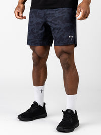 Close-up of a person wearing Essential Training Shorts in dark camo with a small cross logo near the hem, paired with white crew socks featuring a cross and black athletic shoes.