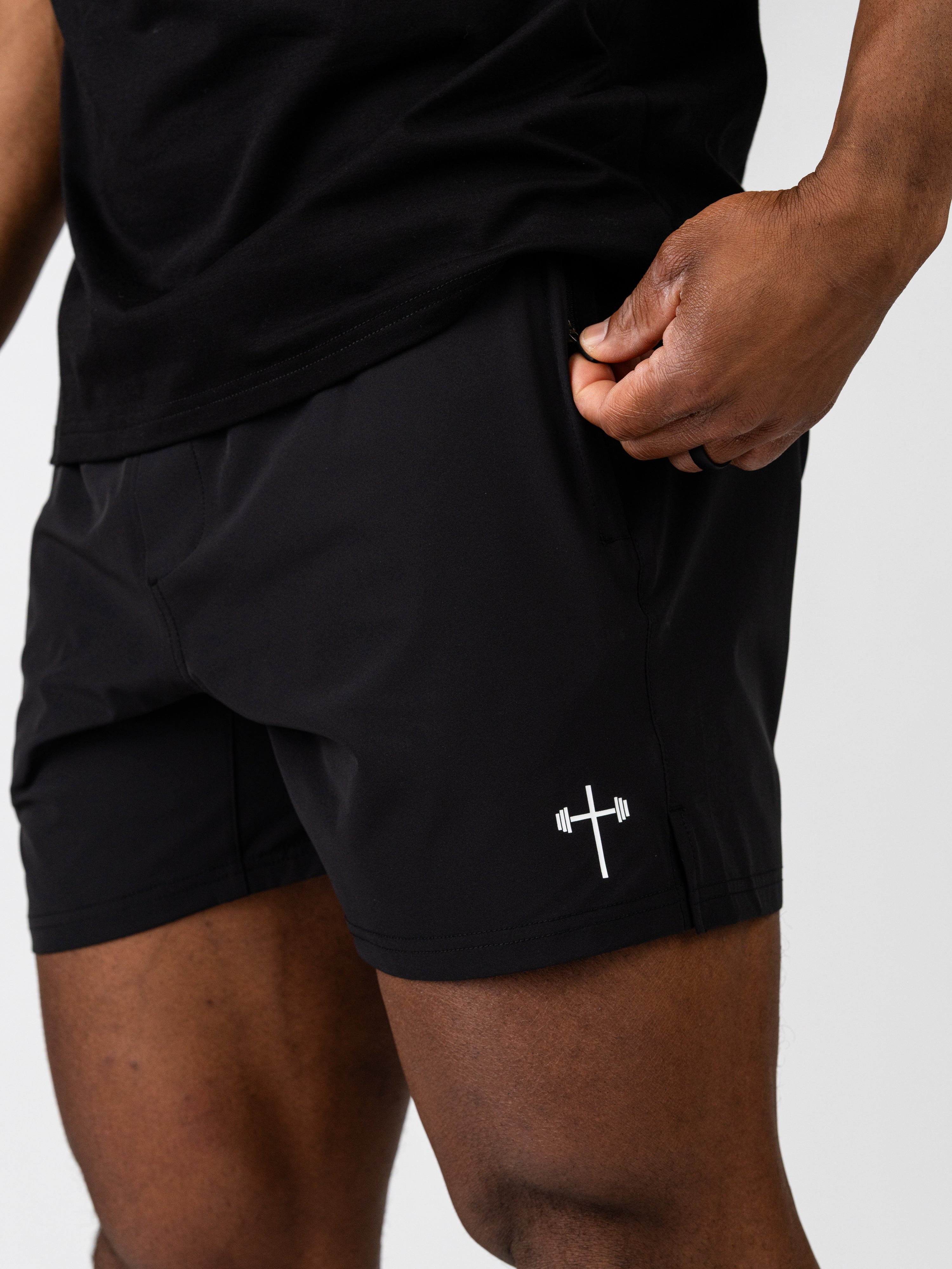 Close-up of a person wearing black Essential Training Shorts with a white Christian barbell cross logo on the left leg, showing the lightweight and breathable fabric designed for active movement.