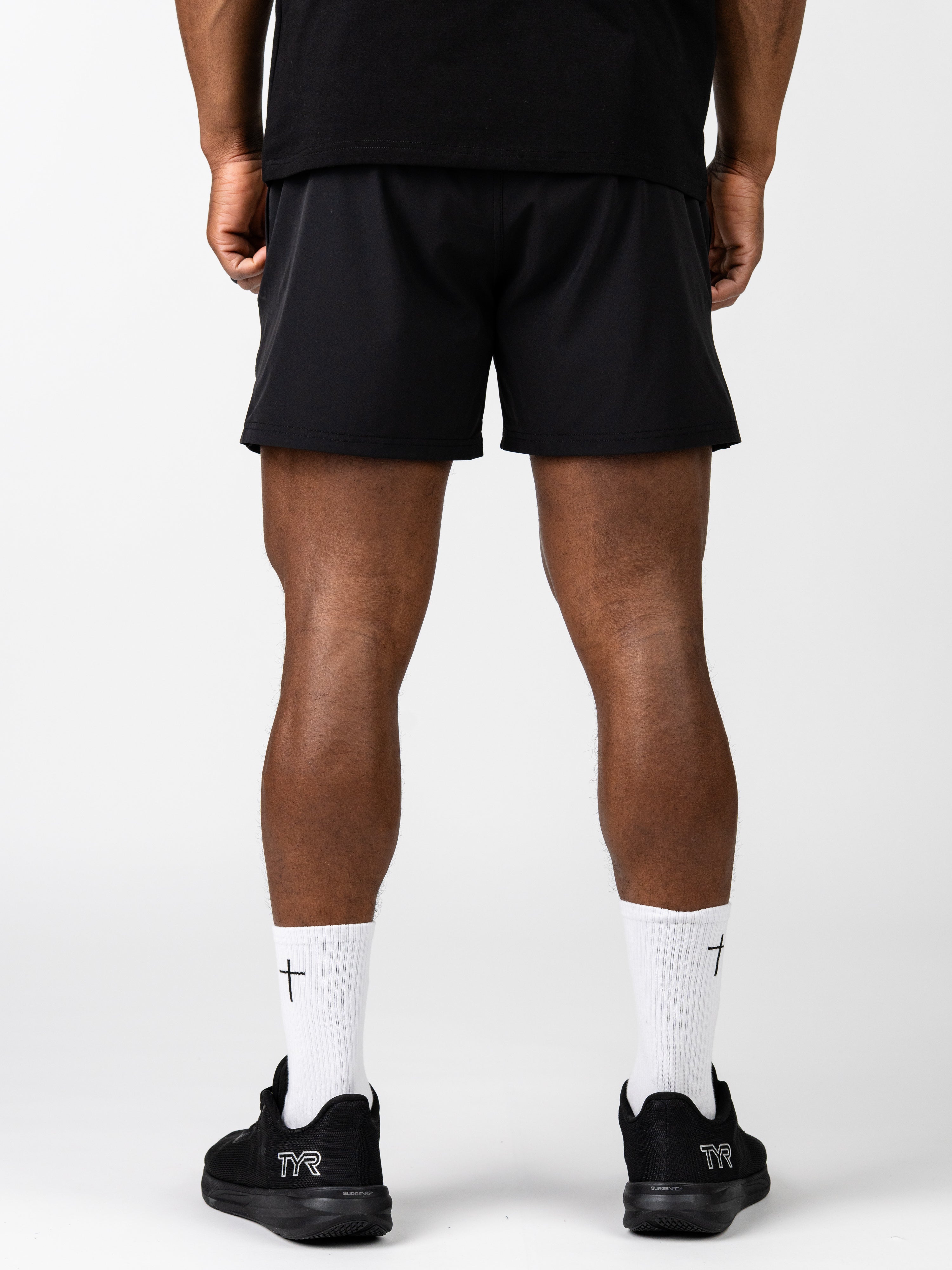 Rear view of a person wearing black Essential Training Shorts with white socks featuring a small cross, paired with black athletic shoes, highlighting lightweight and breathable Christian fitness apparel.