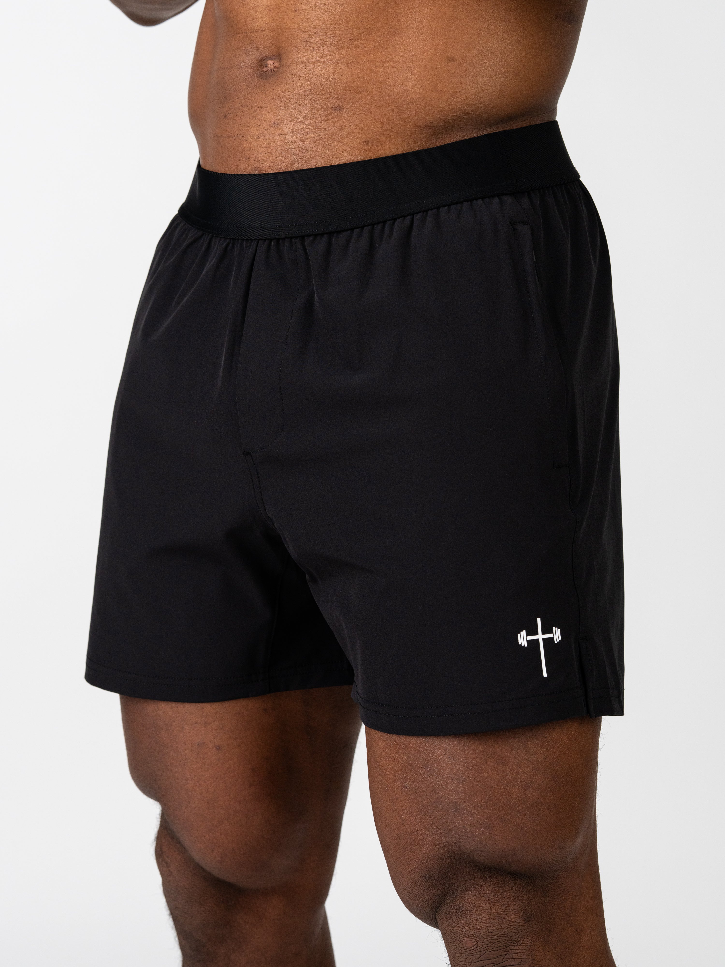Male model wearing black Essential Training Shorts featuring a small white barbell cross logo on the left leg, designed for lightweight, breathable performance during intense workouts.