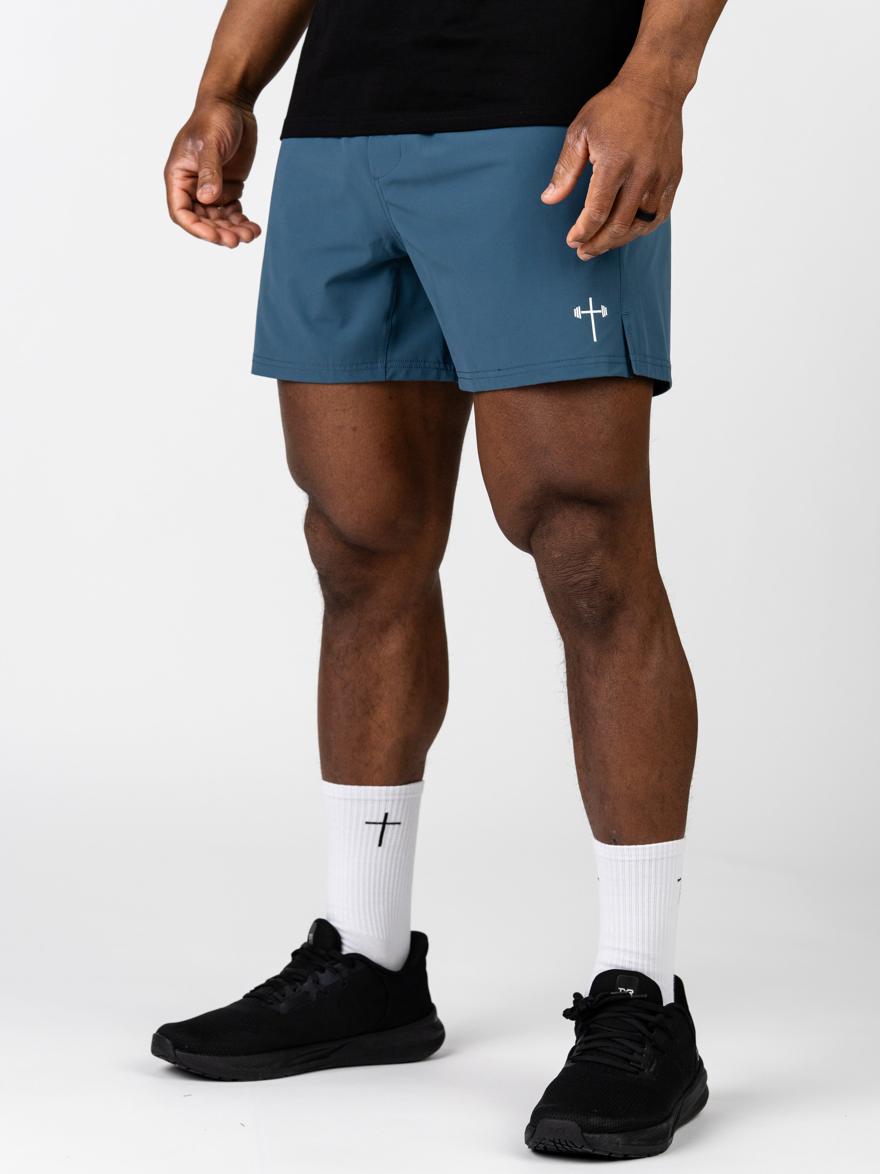 Male model wearing blue Essential Training Shorts with a small white barbell cross logo on the leg, paired with white socks featuring a black cross and black athletic shoes.