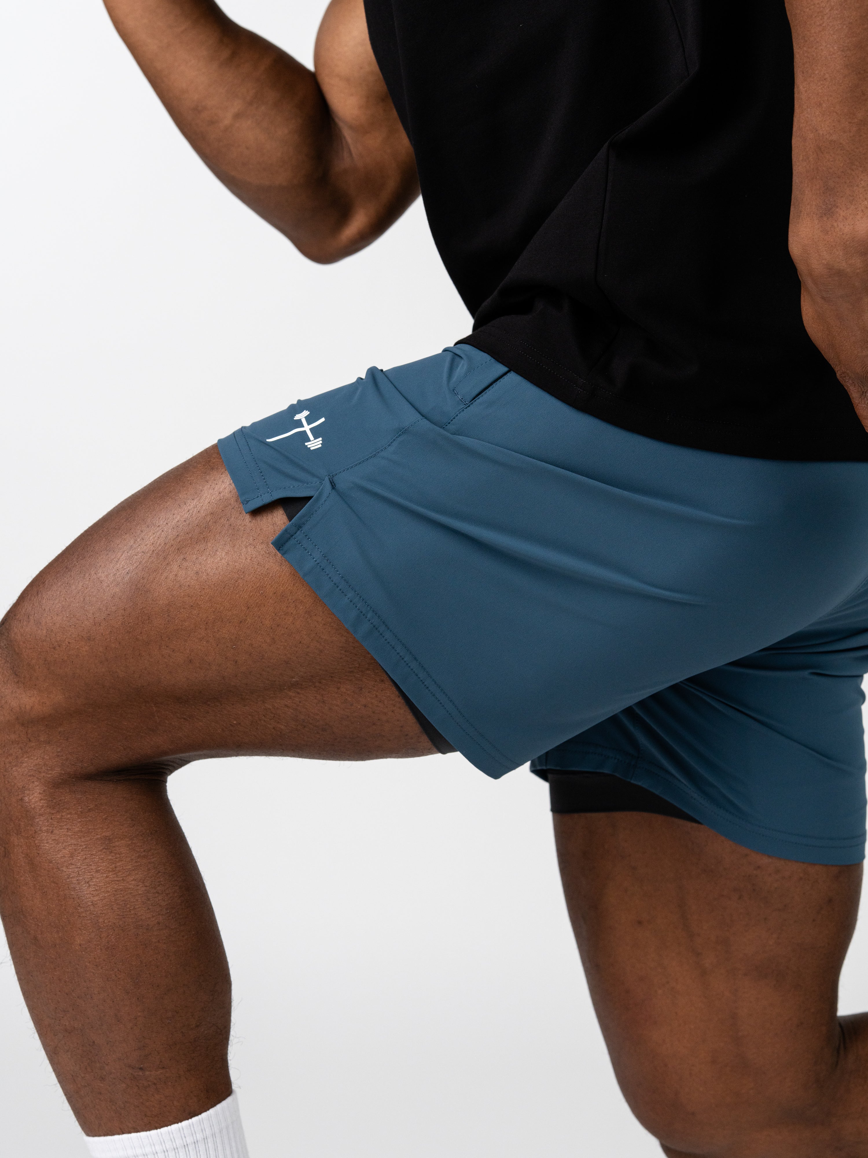 Close-up of a person wearing blue Essential Training Shorts with a small white barbell cross logo on the side, designed for movement and comfort during workouts.