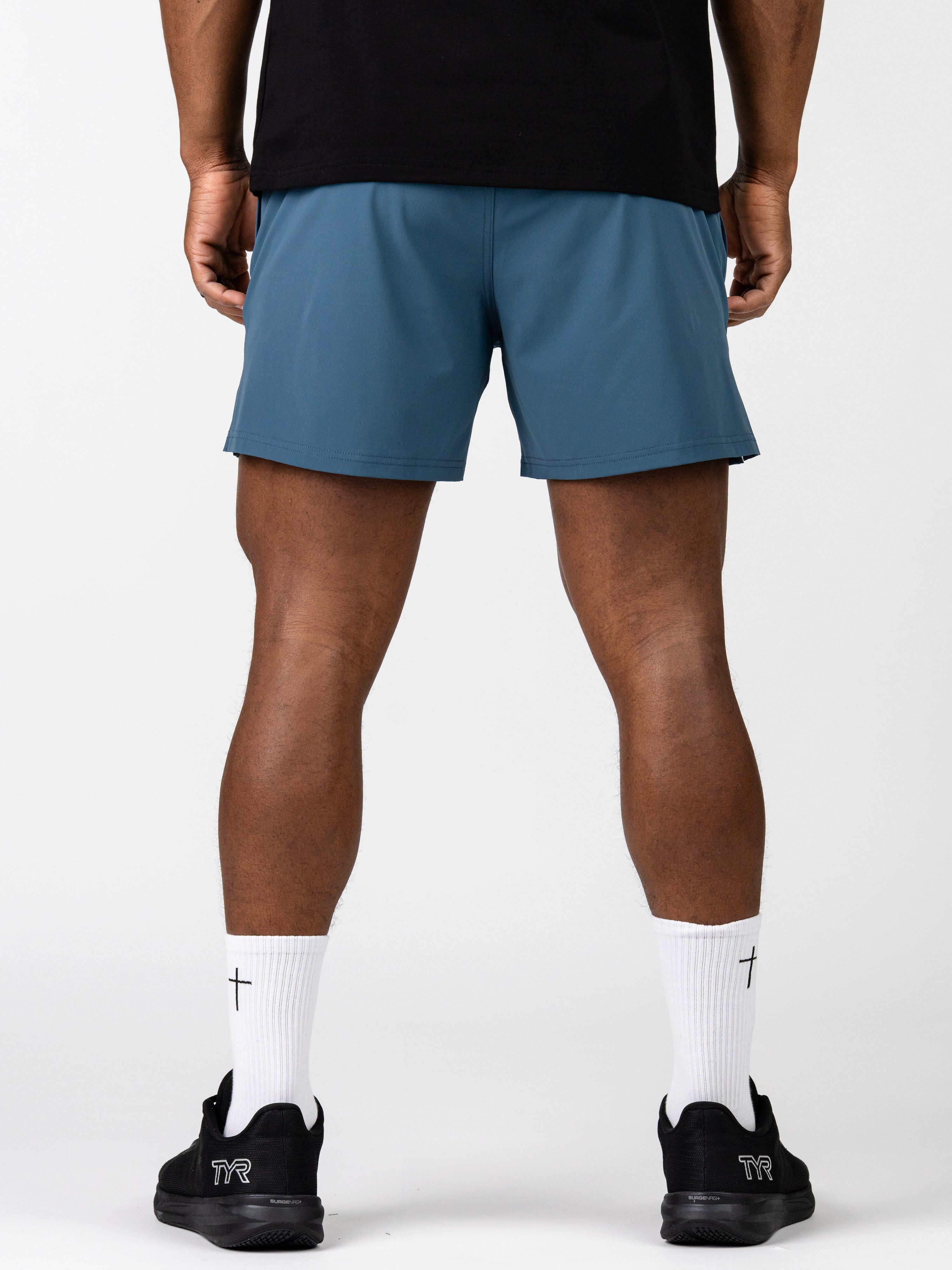 Rear view of a person wearing blue Essential Training Shorts paired with white crew socks featuring a small cross design and black athletic shoes, showcasing lightweight, breathable Christian fitness apparel.