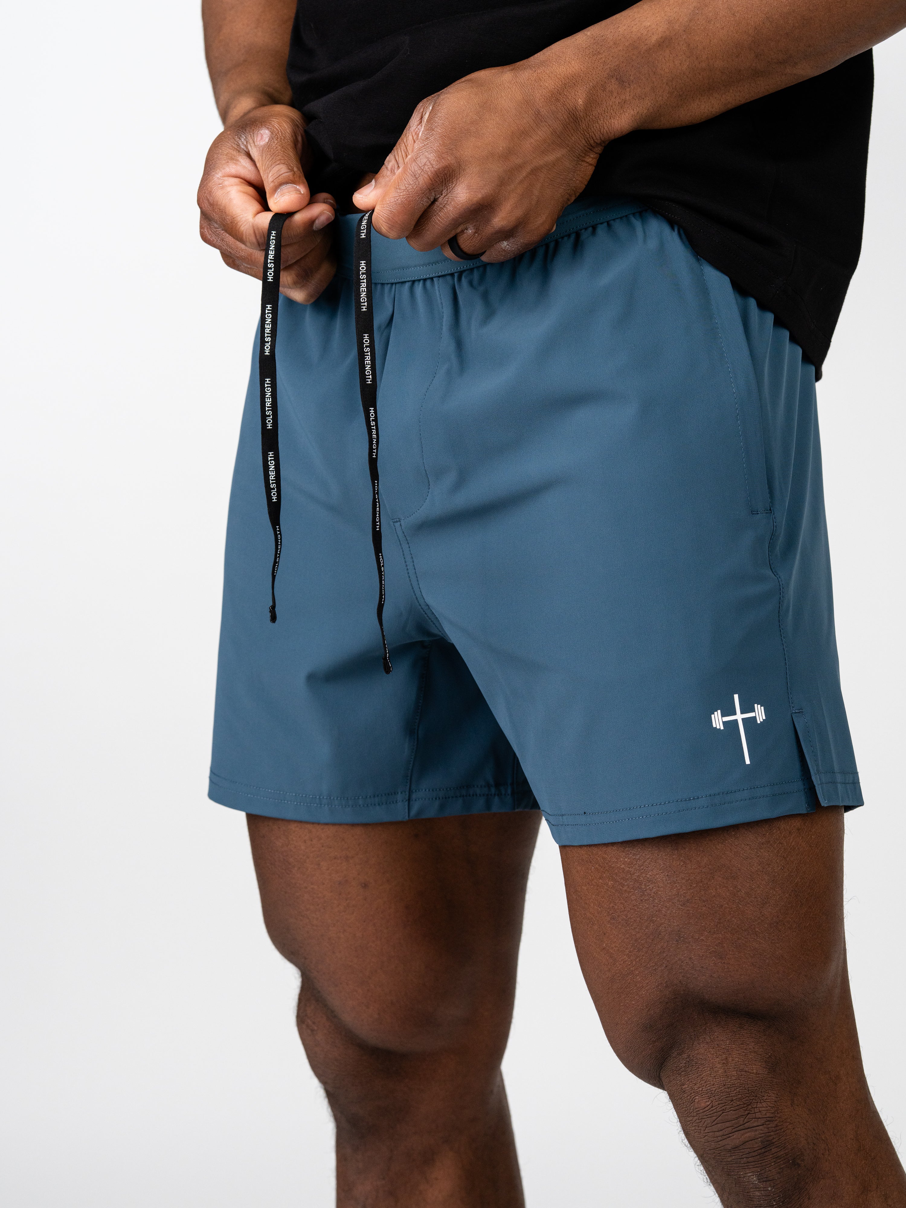 Close-up of a person adjusting the drawstring on blue Essential Training Shorts featuring a small barbell cross logo near the hem, designed for movement and breathability.
