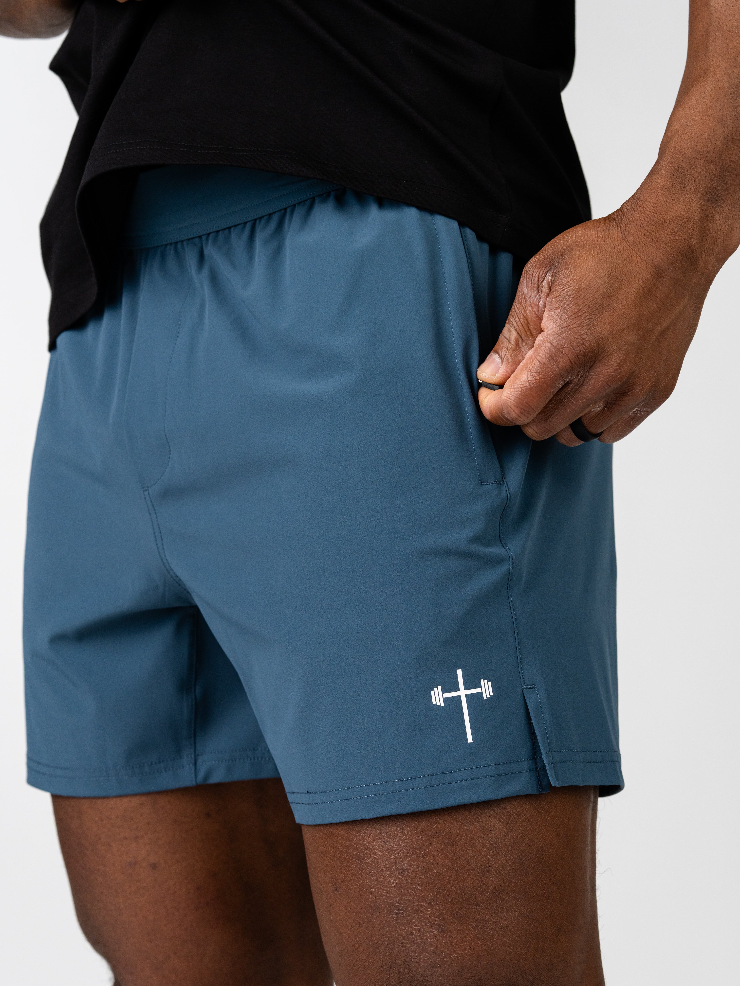 Close-up of a person wearing blue Essential Training Shorts with a small white cross and barbell logo, showcasing lightweight, breathable Christian fitness apparel.