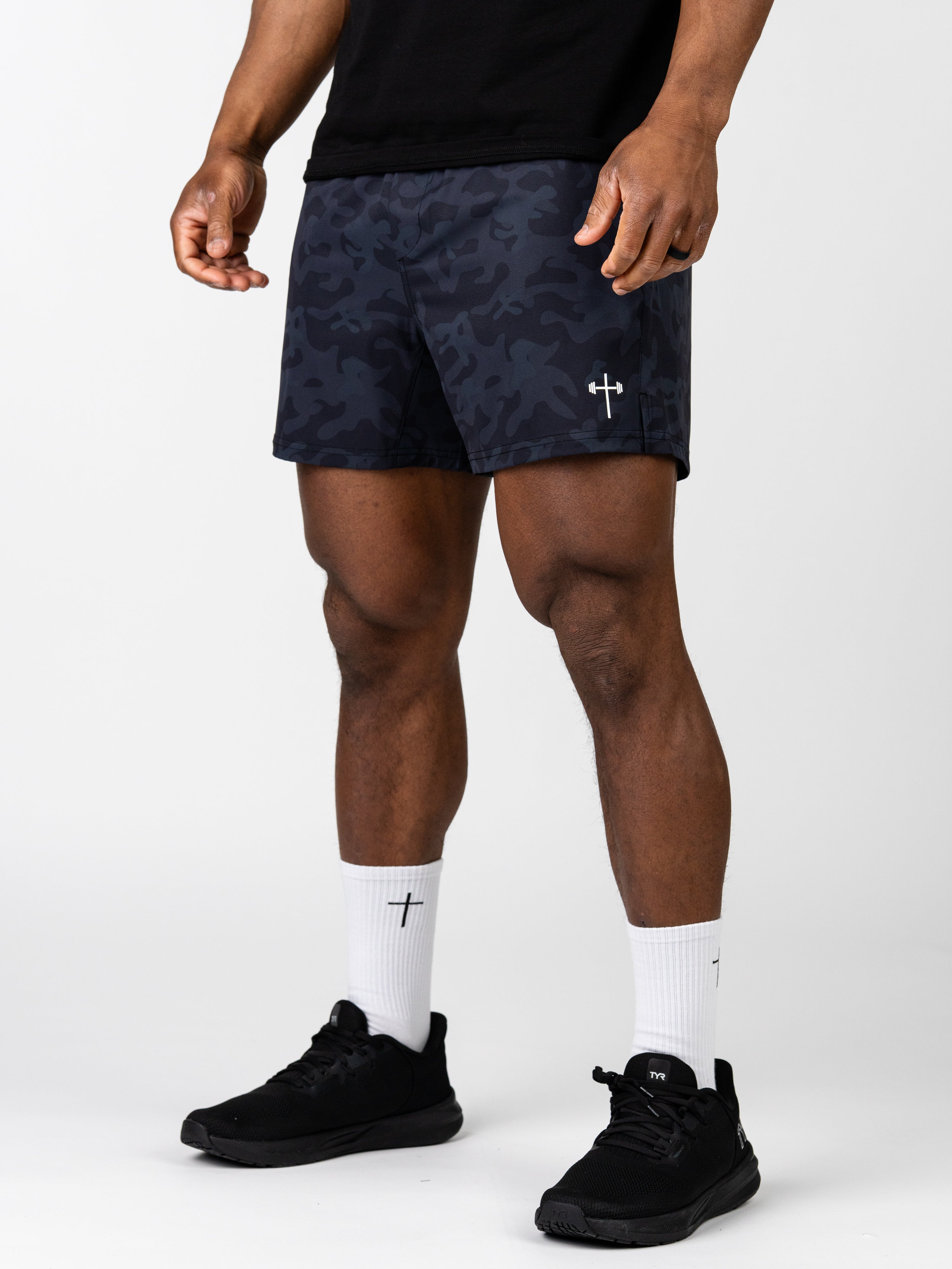 Athlete wearing black camo Essential Training Shorts featuring a subtle Christian cross logo, paired with white socks and black athletic shoes, standing against a plain background.
