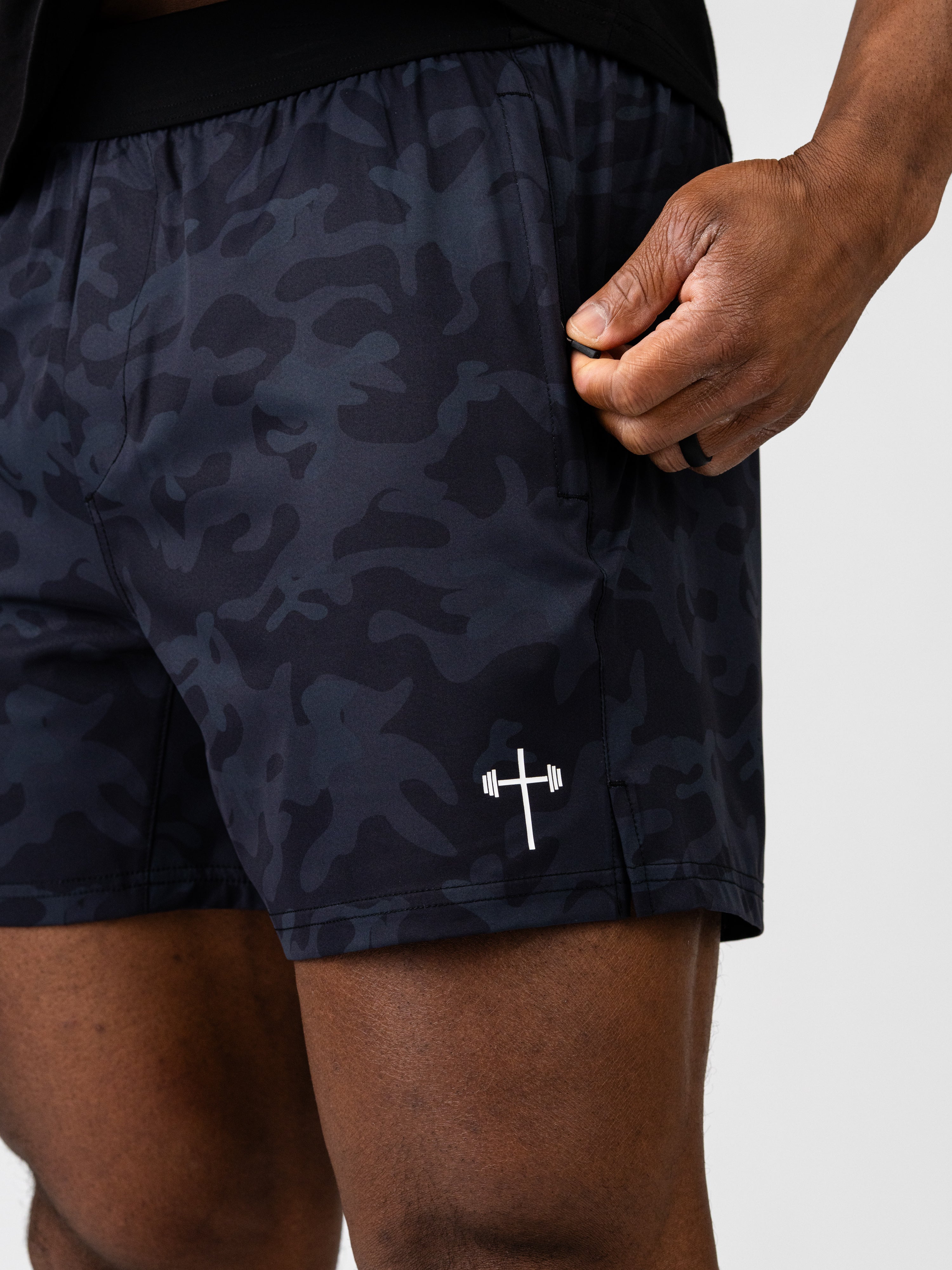 Close-up of a person wearing Essential Training Shorts with a dark camo pattern and a white barbell cross logo on the lower thigh, demonstrating a lightweight and flexible fit.