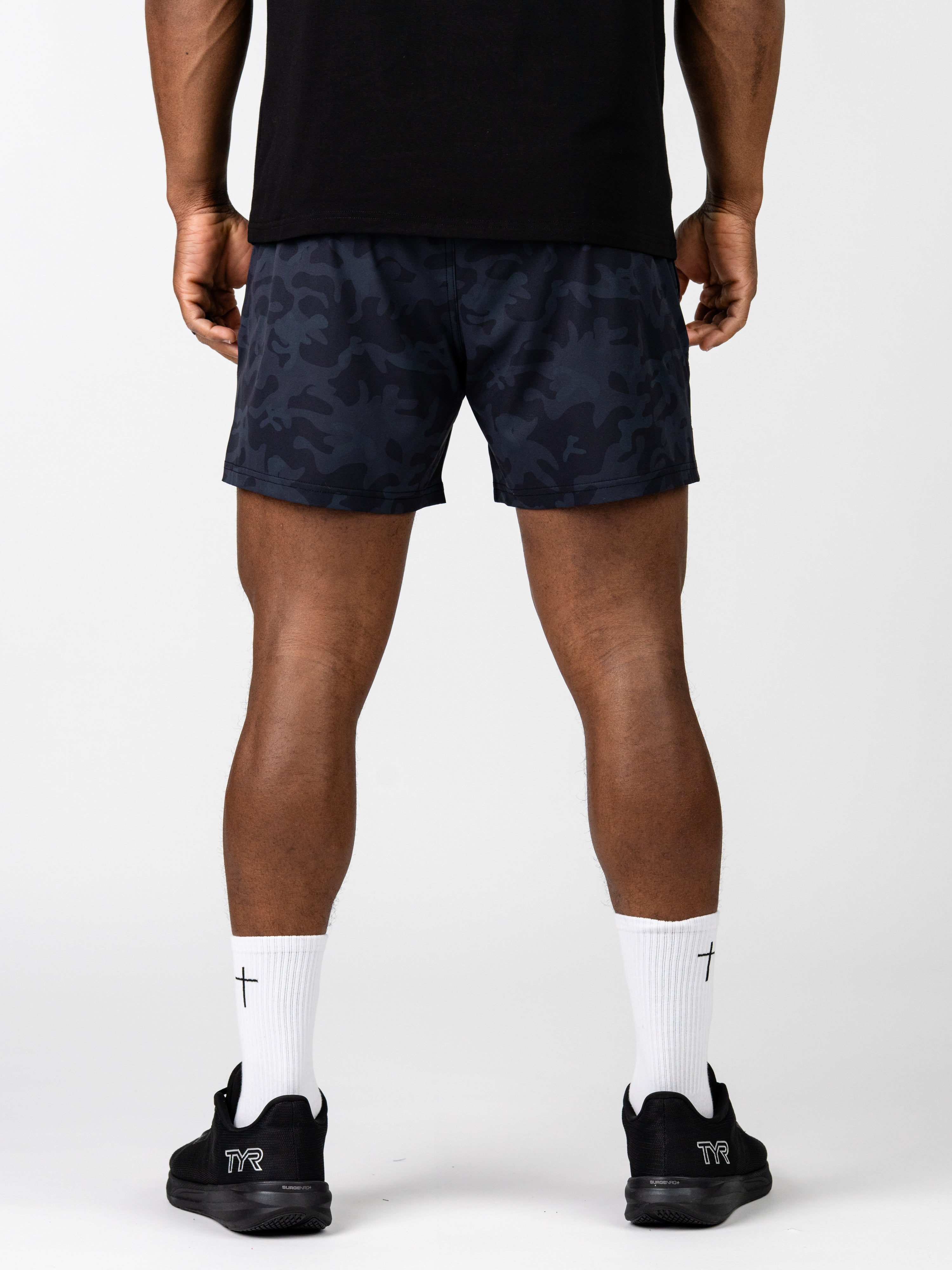 Rear view of a person wearing Essential Training Shorts in a dark camo pattern paired with white crew socks featuring a small cross, and black athletic shoes.