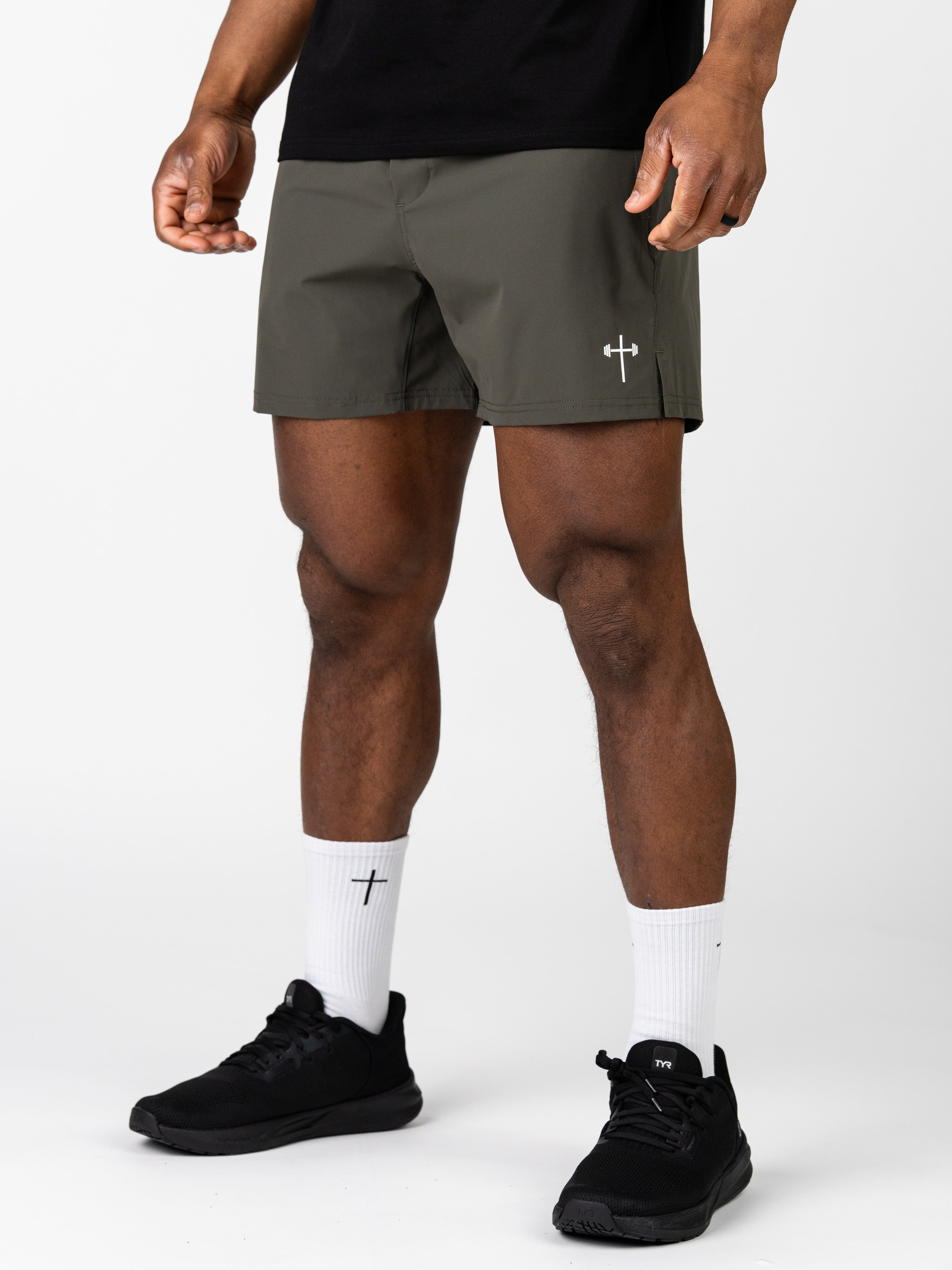 Man wearing Essential Training Shorts in olive green paired with white crew socks and black athletic shoes, featuring a subtle Christian barbell cross logo on the shorts.