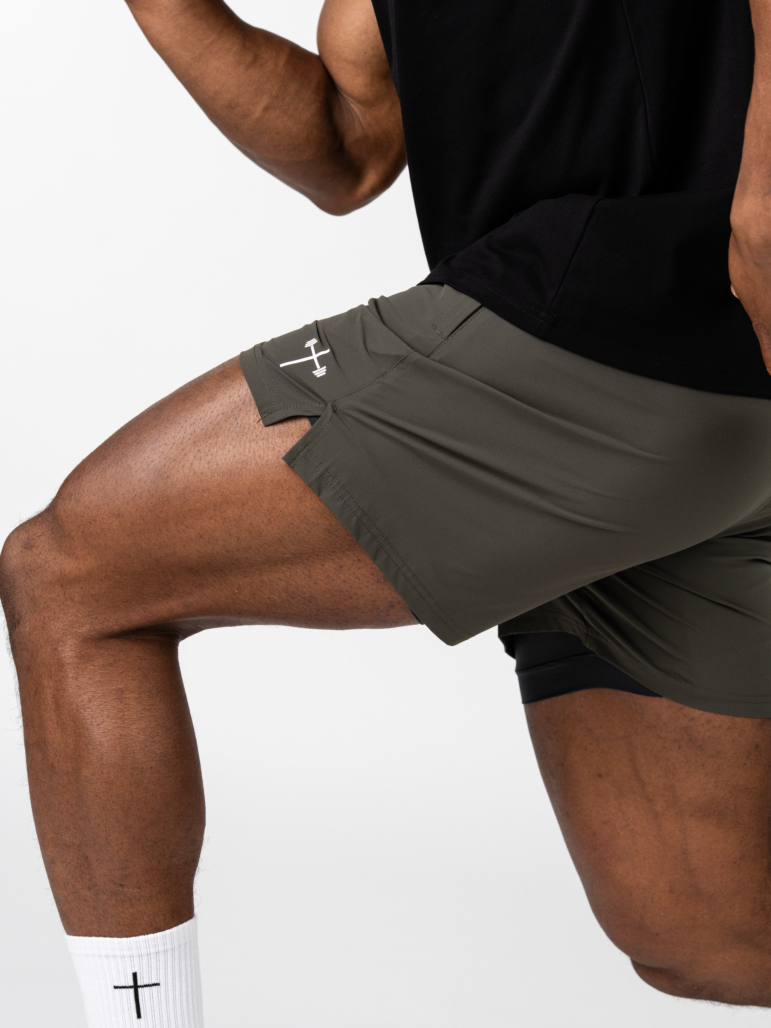 A close-up of a person wearing olive green Essential Training Shorts with a white Christian cross and barbell logo, demonstrating flexibility in a workout pose.